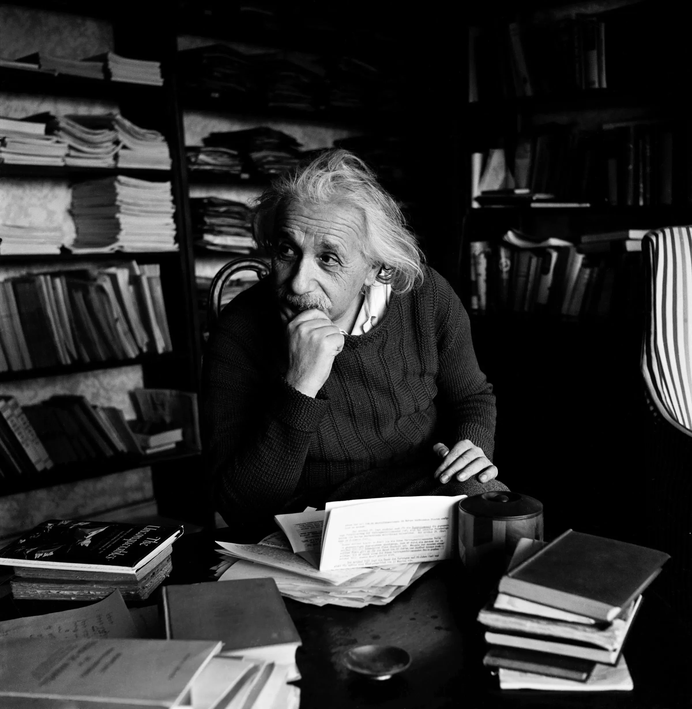 A black and white photo of Albert Einstein sitting at a cluttered desk with books, papers, and a coffee cup, with shelves full of stacks of books behind him.
