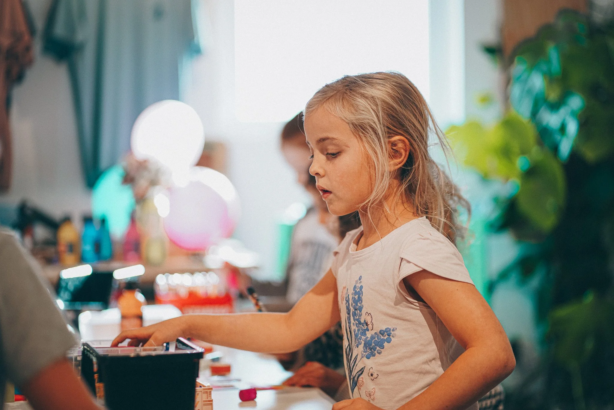 A young girl with blonde hair and a white shirt playing with toys at a table indoors, with other children around and colorful balloons in the background.