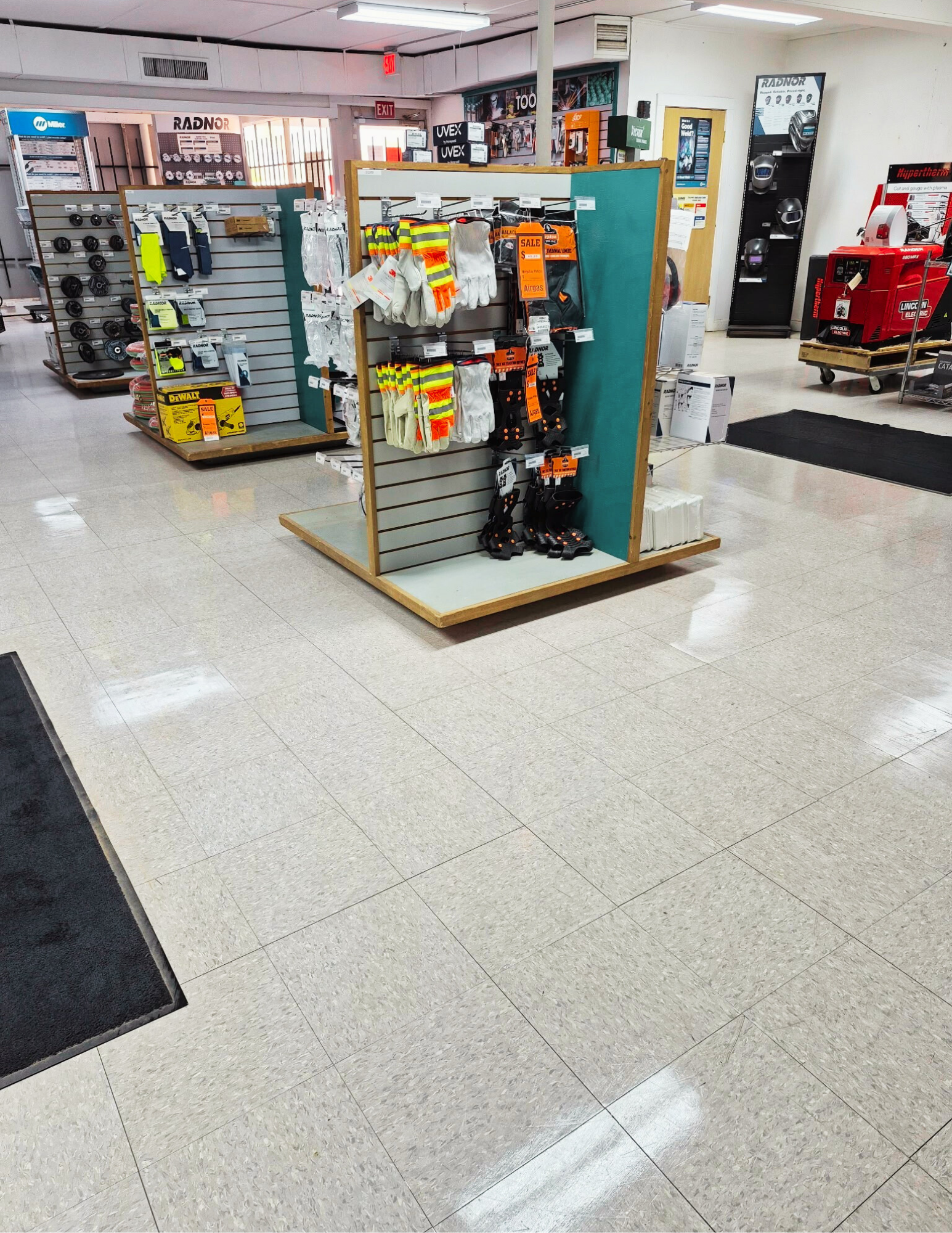 Interior of a store with display racks holding safety gloves, gloves, and knee pads. Various equipment and tools are visible in the background, including a red machine on a pallet.