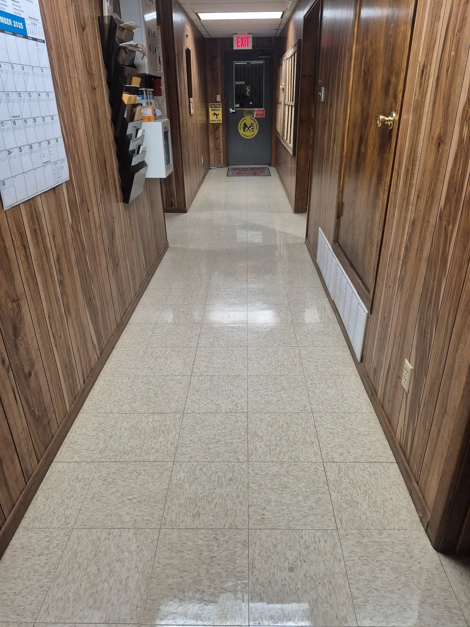 A corridor with wood-paneled walls, tiled floor, and a door at the end with an illuminated exit sign above it.
