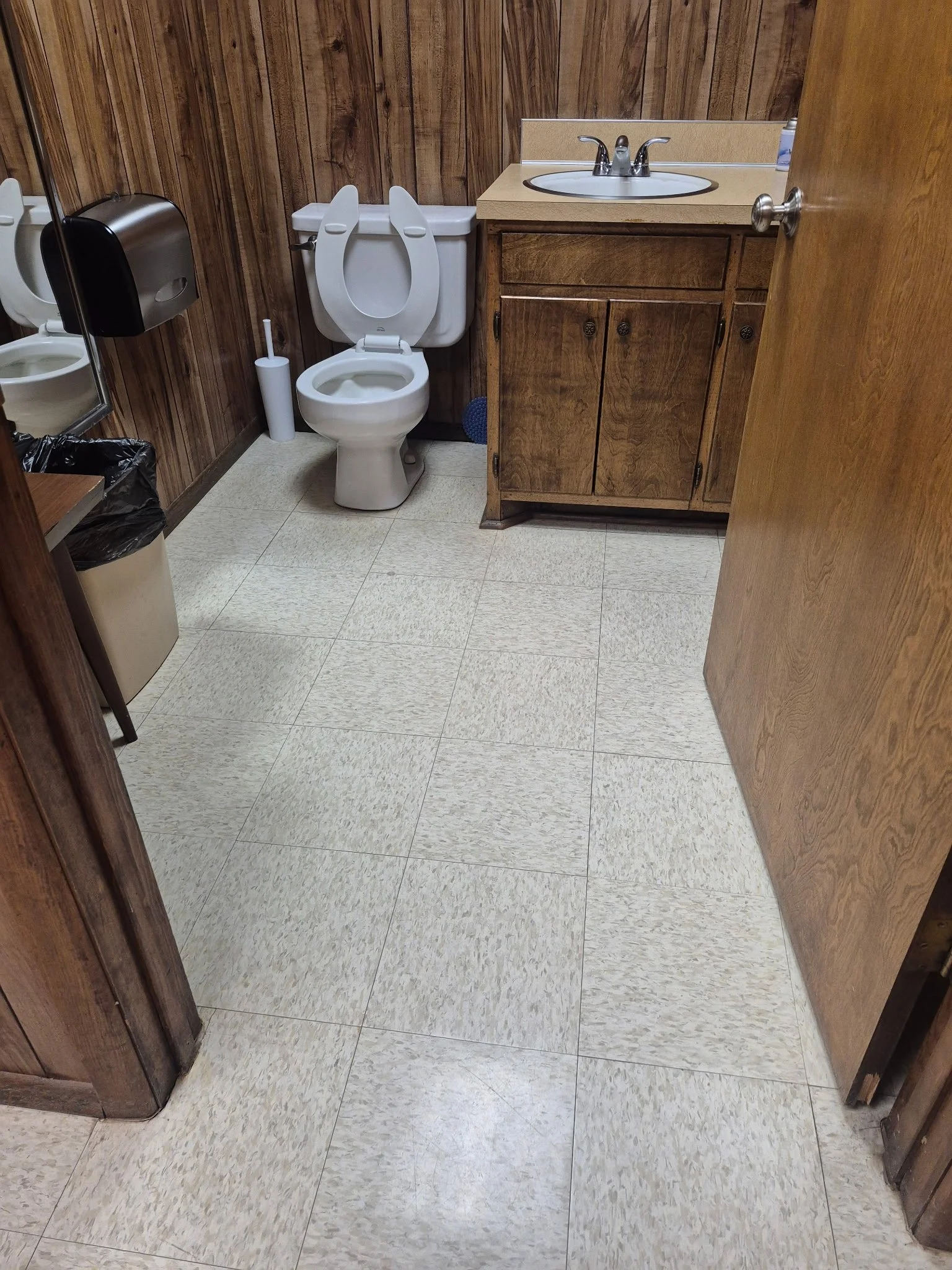 E&E Solutions Cleaning A small bathroom with wood panel walls, a toilet with the lid up, a wooden vanity with a sink, a mirror, a toilet paper holder, a trash can, and a small table.