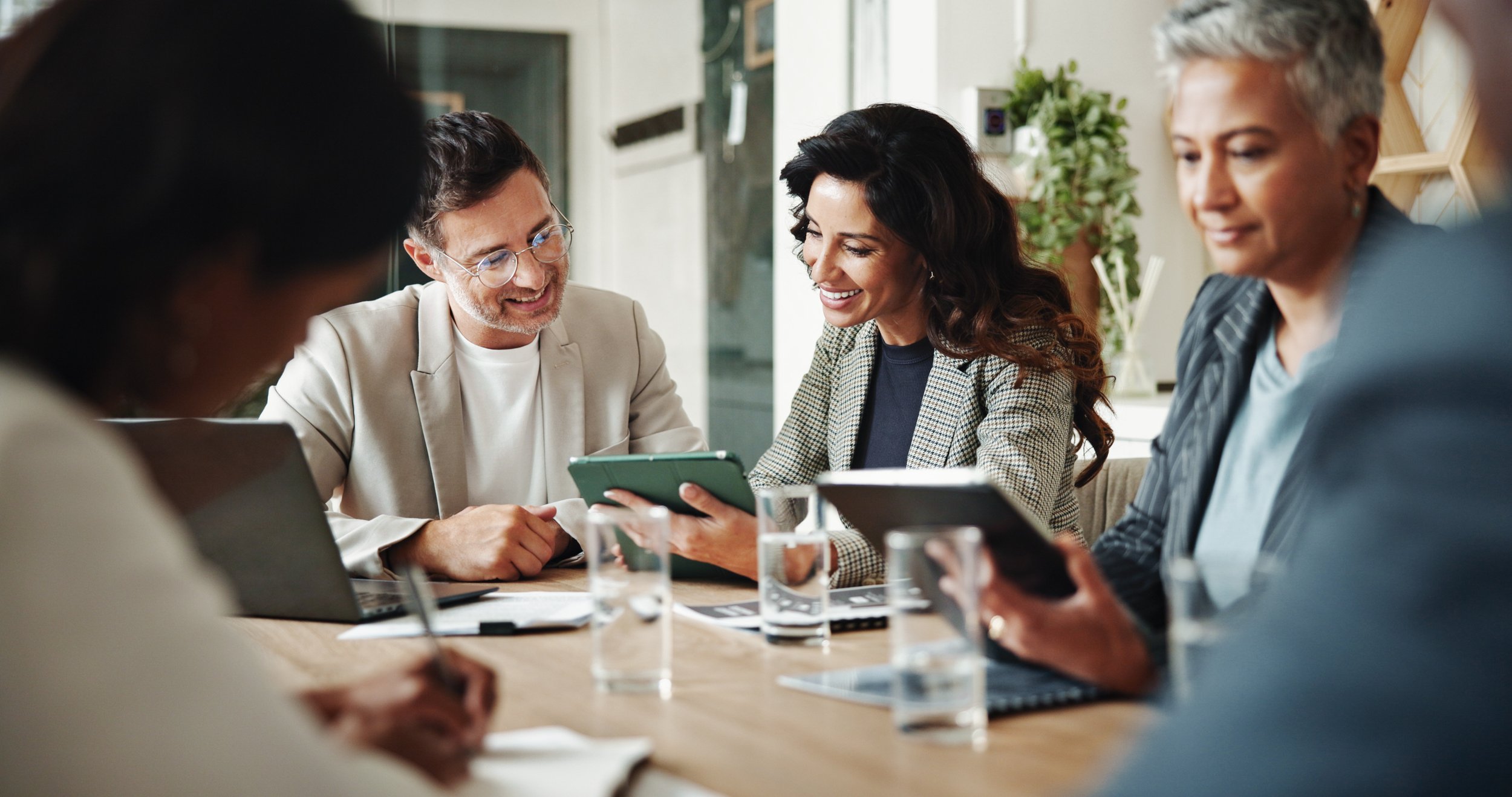 Group of five diverse professionals sitting at a table in a meeting, smiling and engaging with each other, with laptops and tablets.