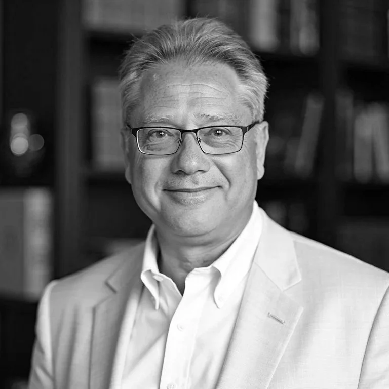 Portrait of a middle-aged man with gray hair and glasses, wearing a cream-colored blazer and white shirt, standing in front of a bookshelf.