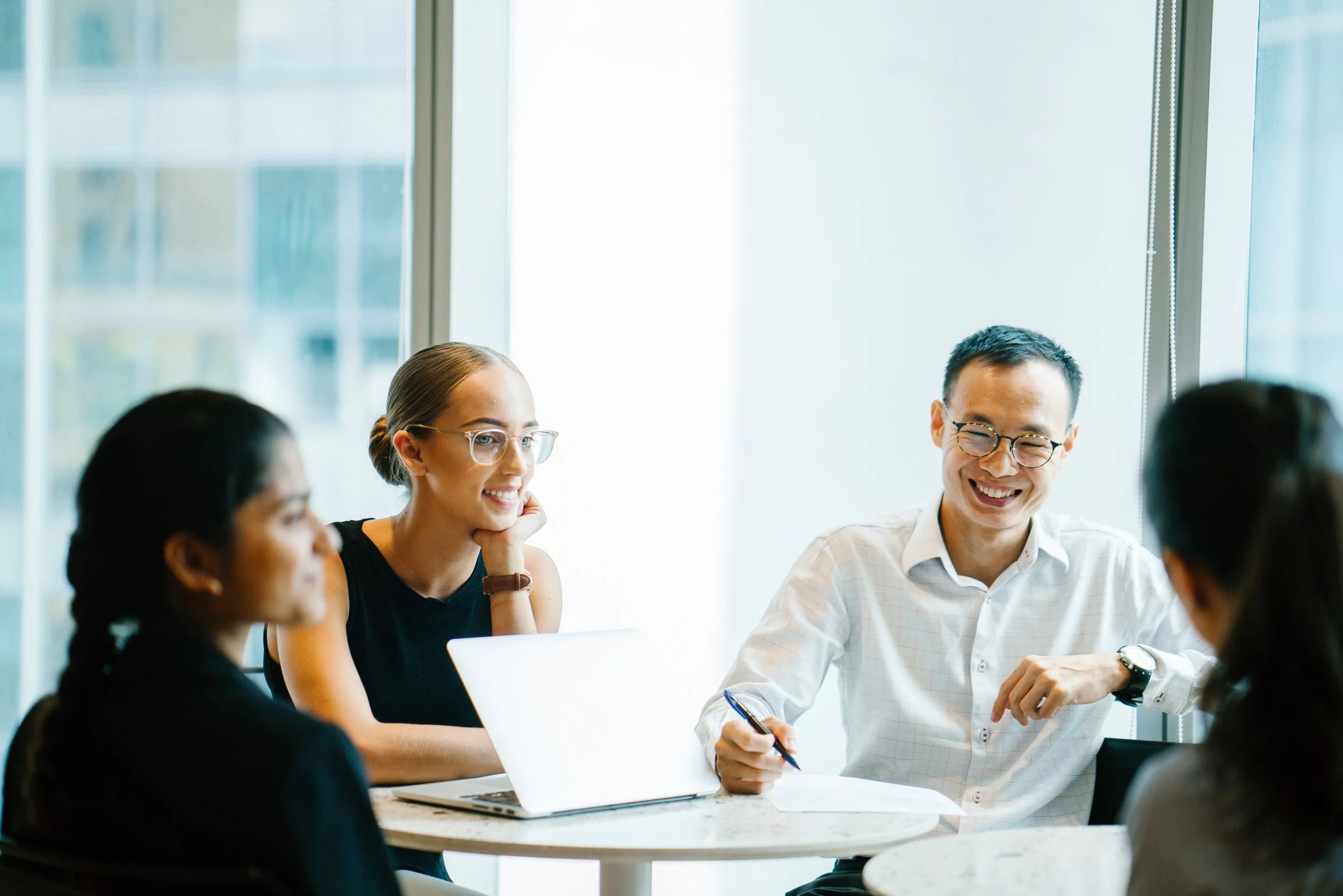 Business team having a meeting in a bright, modern office, smiling and discussing around a table with laptops.