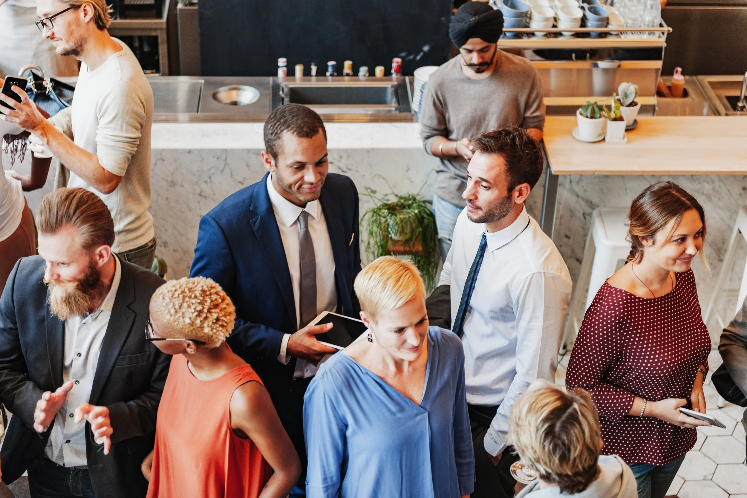 Group of diverse people socializing at a cafe or restaurant, some holding drinks, others using smartphones, in a modern setting with a marble countertop and potted plants.