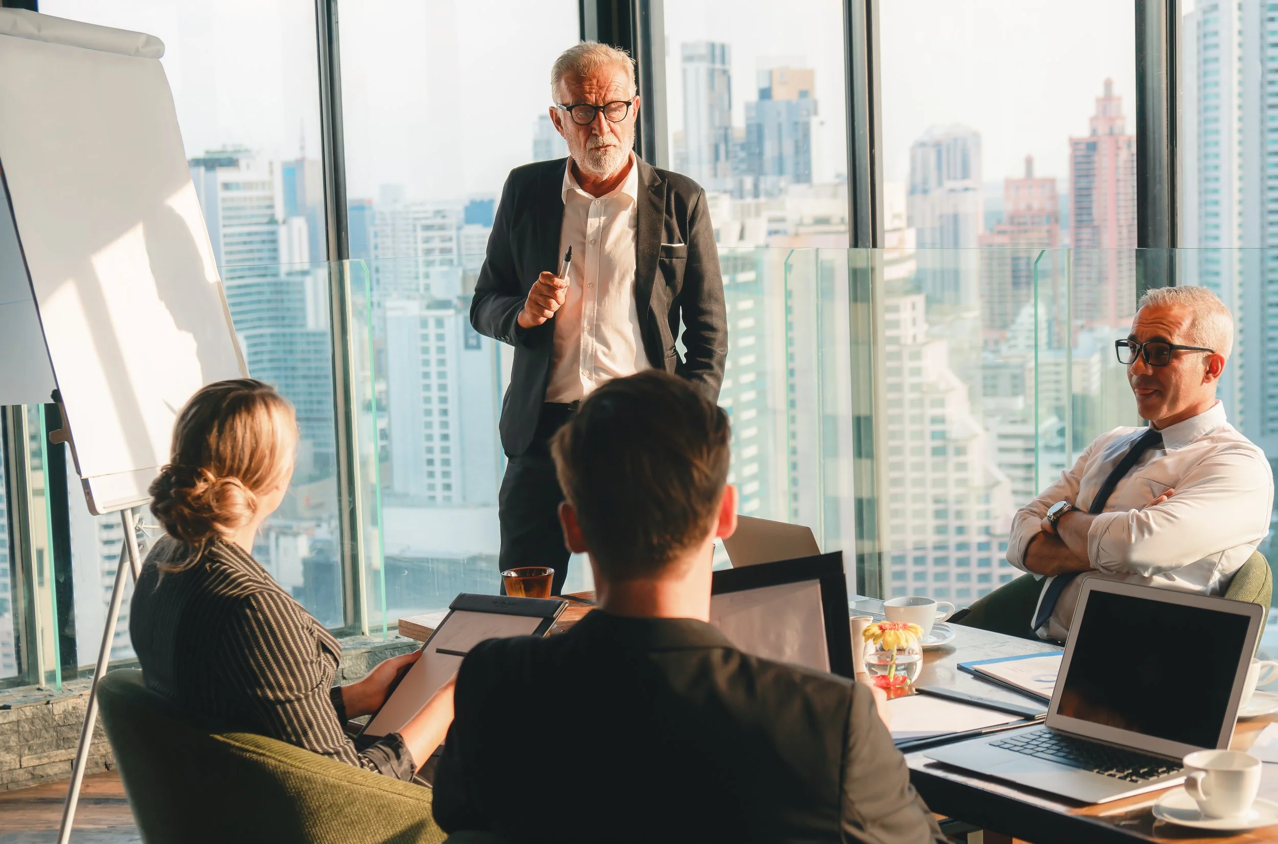 Business meeting in a high-rise office with a presenter standing near a whiteboard, five people seated around a table with laptops, coffee cups, and notebooks, with a city skyline visible through floor-to-ceiling windows.