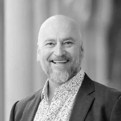 A smiling man with a bald head and gray beard, wearing a dark blazer and a floral shirt, standing outdoors with a blurred background.
