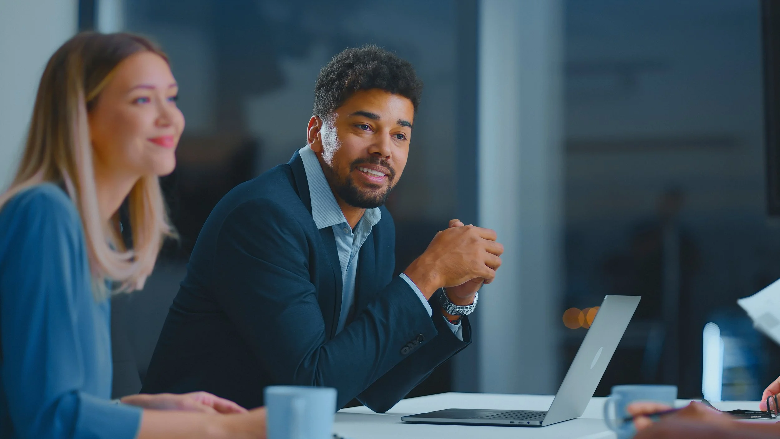 Two business professionals, a woman and a man, sitting at a conference table discussing in an office setting, with a laptop and coffee mugs present.