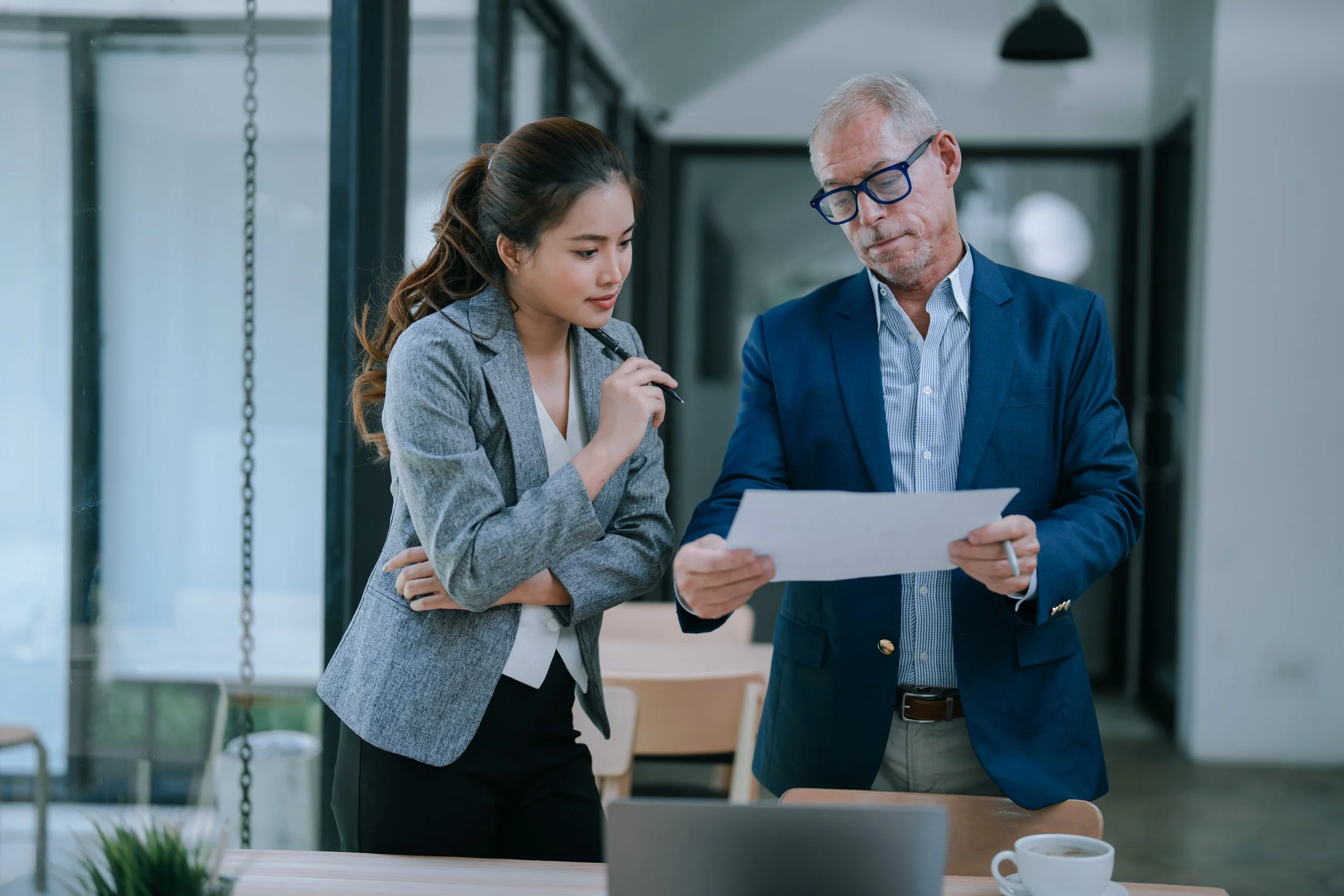 A man and woman in business attire standing at a table in an office, reviewing a document together.