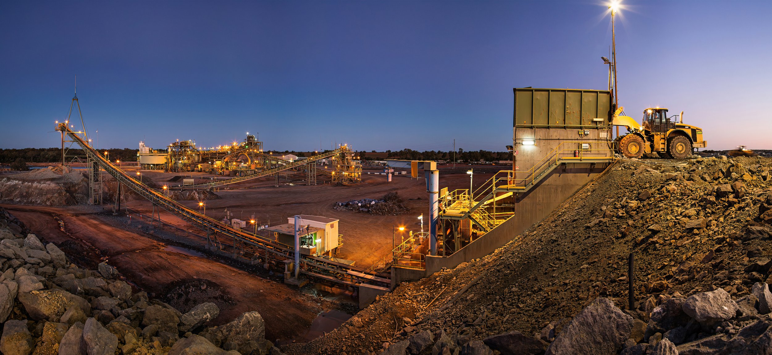 Nighttime view of an industrial mining site with a large excavator on a mound of rocks, conveyor belt, and multiple illuminated factories and processing equipment in the background.