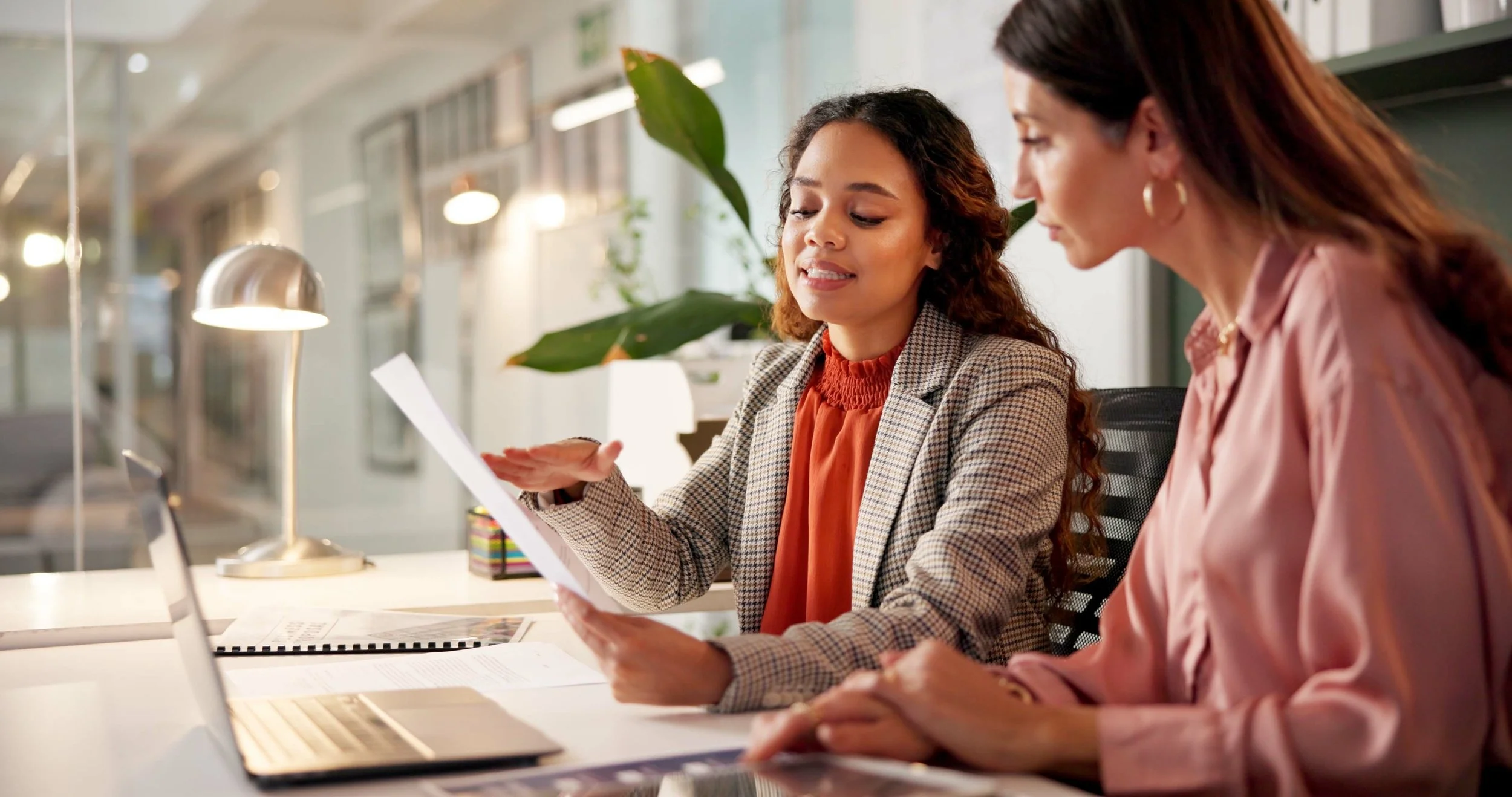 Two women sitting at a desk in a modern office, reviewing documents and working on a laptop.