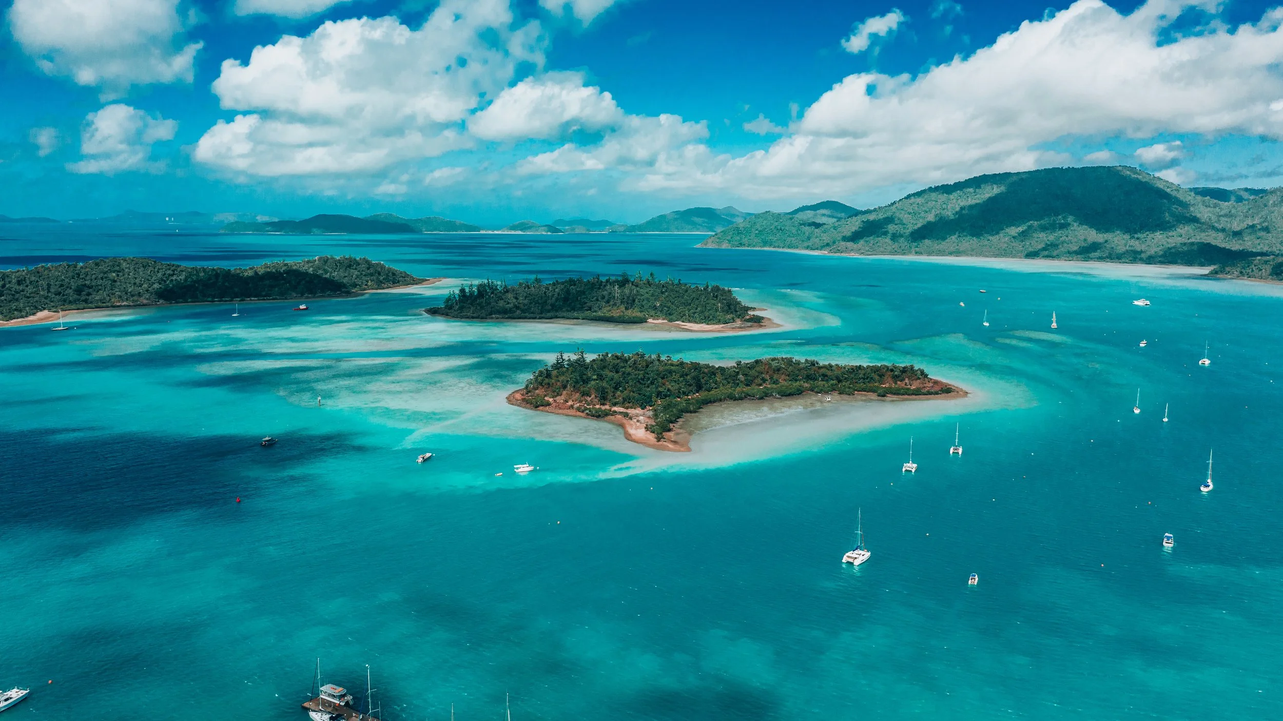 Aerial view of turquoise ocean waters with small islands and sailboats, mountains in the background, under a partly cloudy sky.