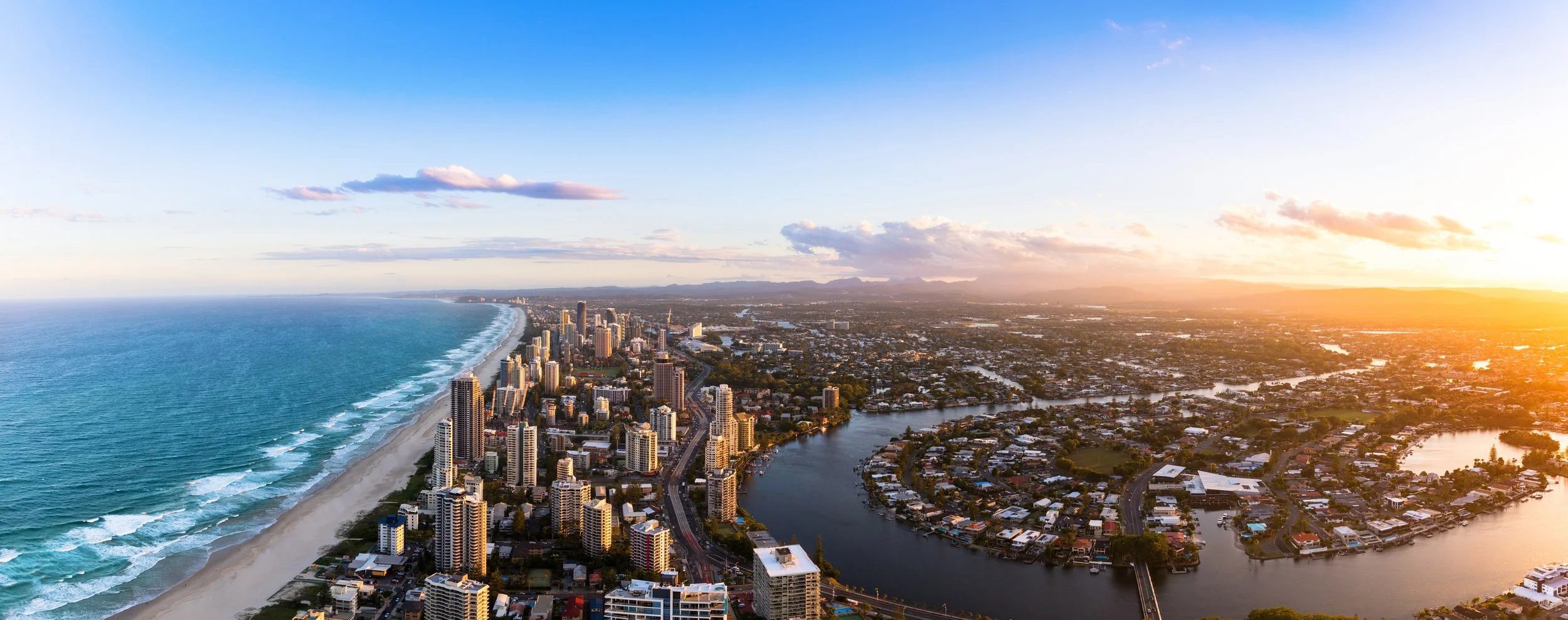 Aerial view of a coastal city with high-rise buildings along the shoreline, a river flowing through the city, and a sunset casting a warm glow over the scene.