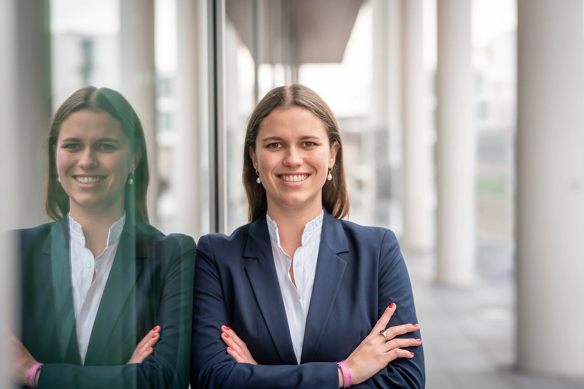 Eine professionelle Frau in Business-Kleidung steht an einem Fenster und lächelt in die Kamera, während sie ihre Arme vor der Brust verschränkt.