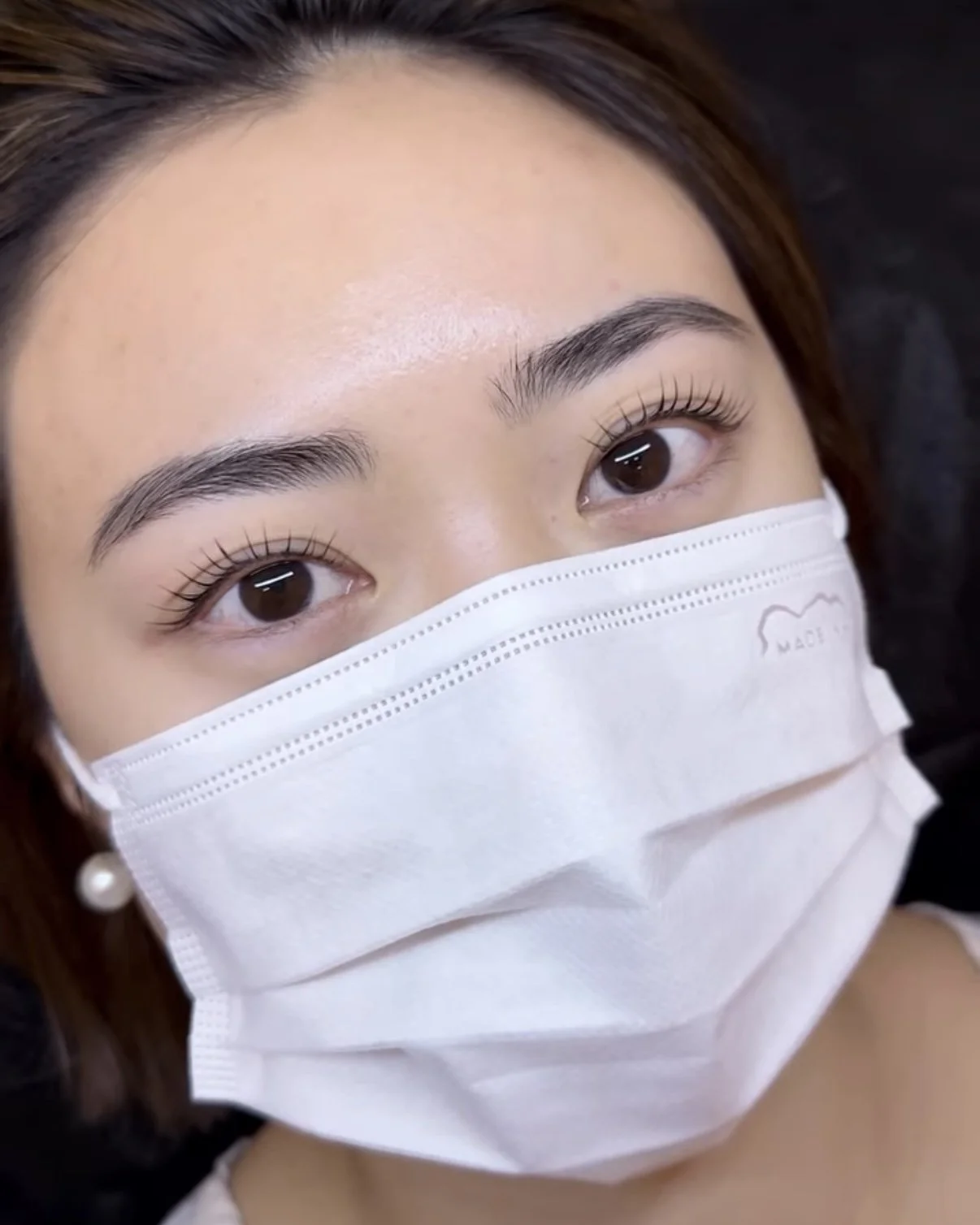 Close-up of a woman with well-groomed eyebrows and long eyelashes wearing a white face mask and pearl earrings.