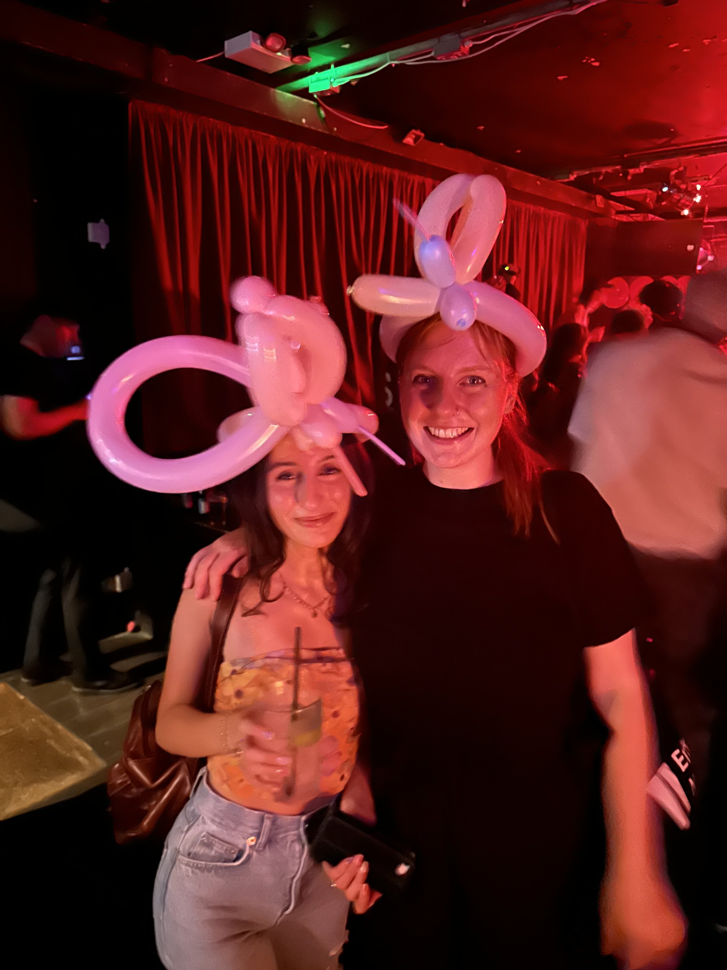Two women at a party wearing balloon hats, one holding a drink, in a dimly lit setting with red lighting.