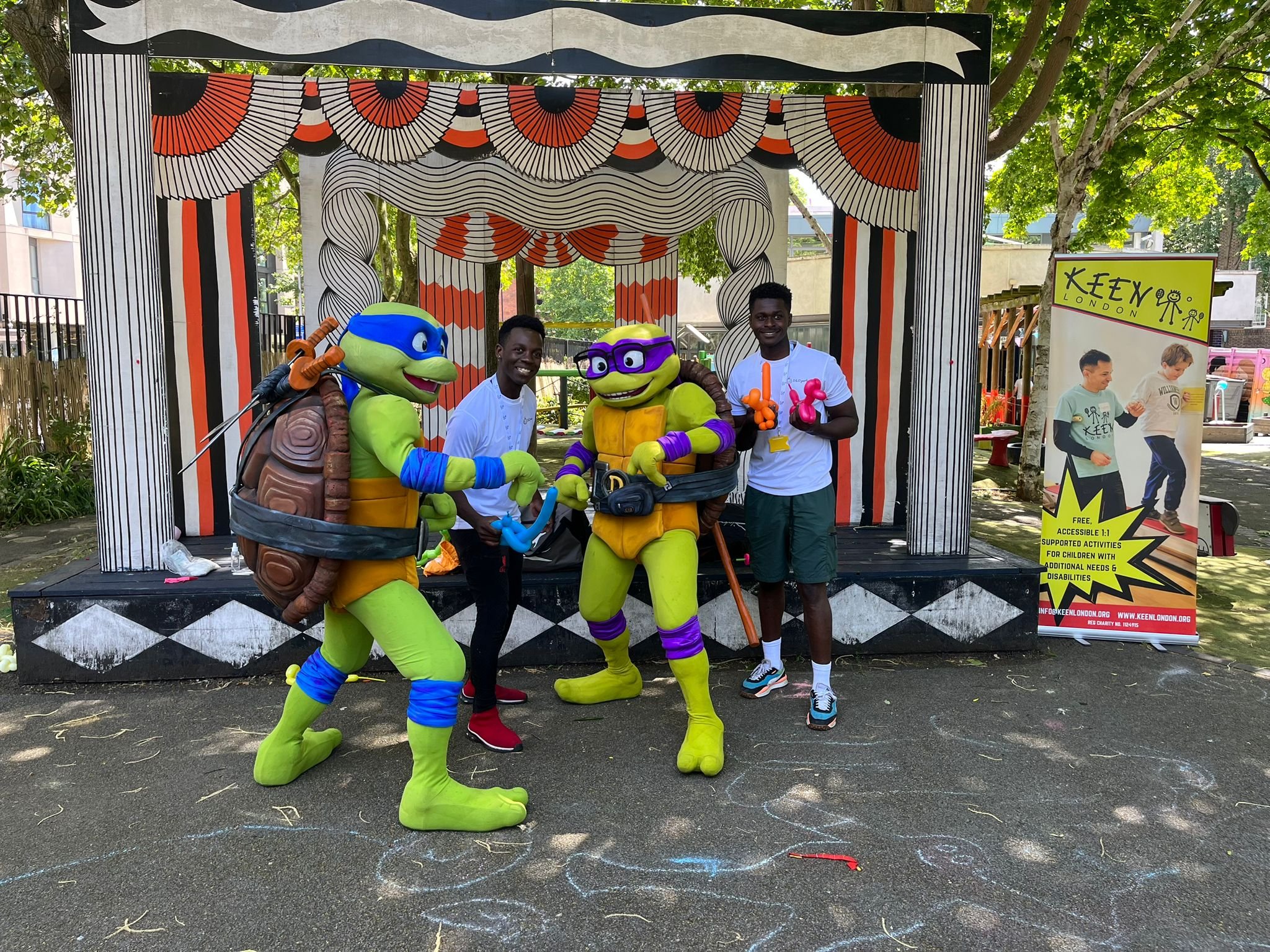Two people posing with characters in turtle costumes in front of a decorated stage in an outdoor setting, with a banner promoting KEEN London activities for children nearby.