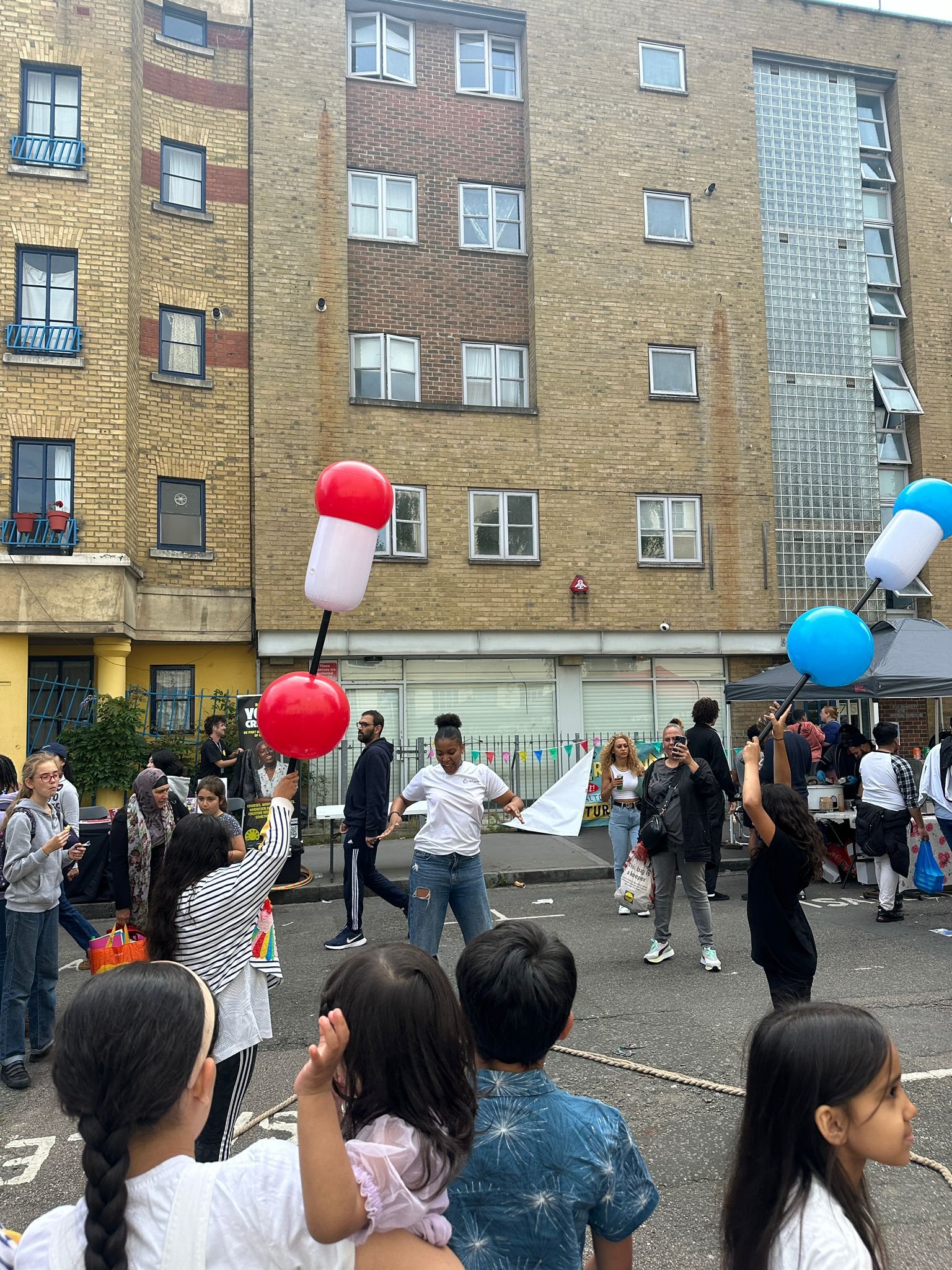 A diverse group of people, including adults and children, gathered outdoors at a street event with colorful decorations. Attendees hold large balloon props shaped like pills in red, white, and blue. The scene is lively, with a multi-story brick apart