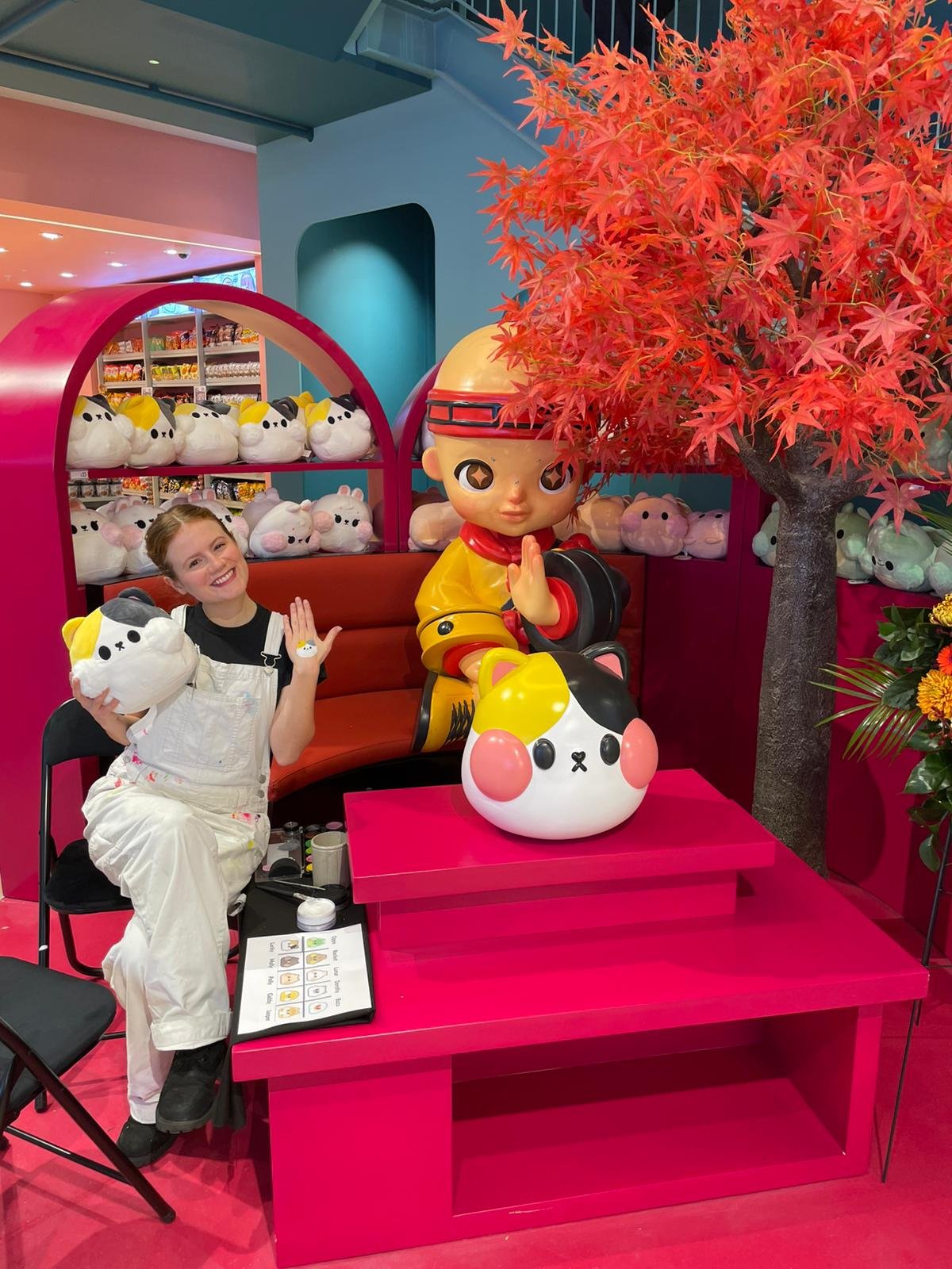 A person sitting next to a large cartoon statue in a vibrant, colorful store setting. The background has shelves filled with plush toys and a large orange-red artificial tree. The person holds a plush toy with smiling expression.