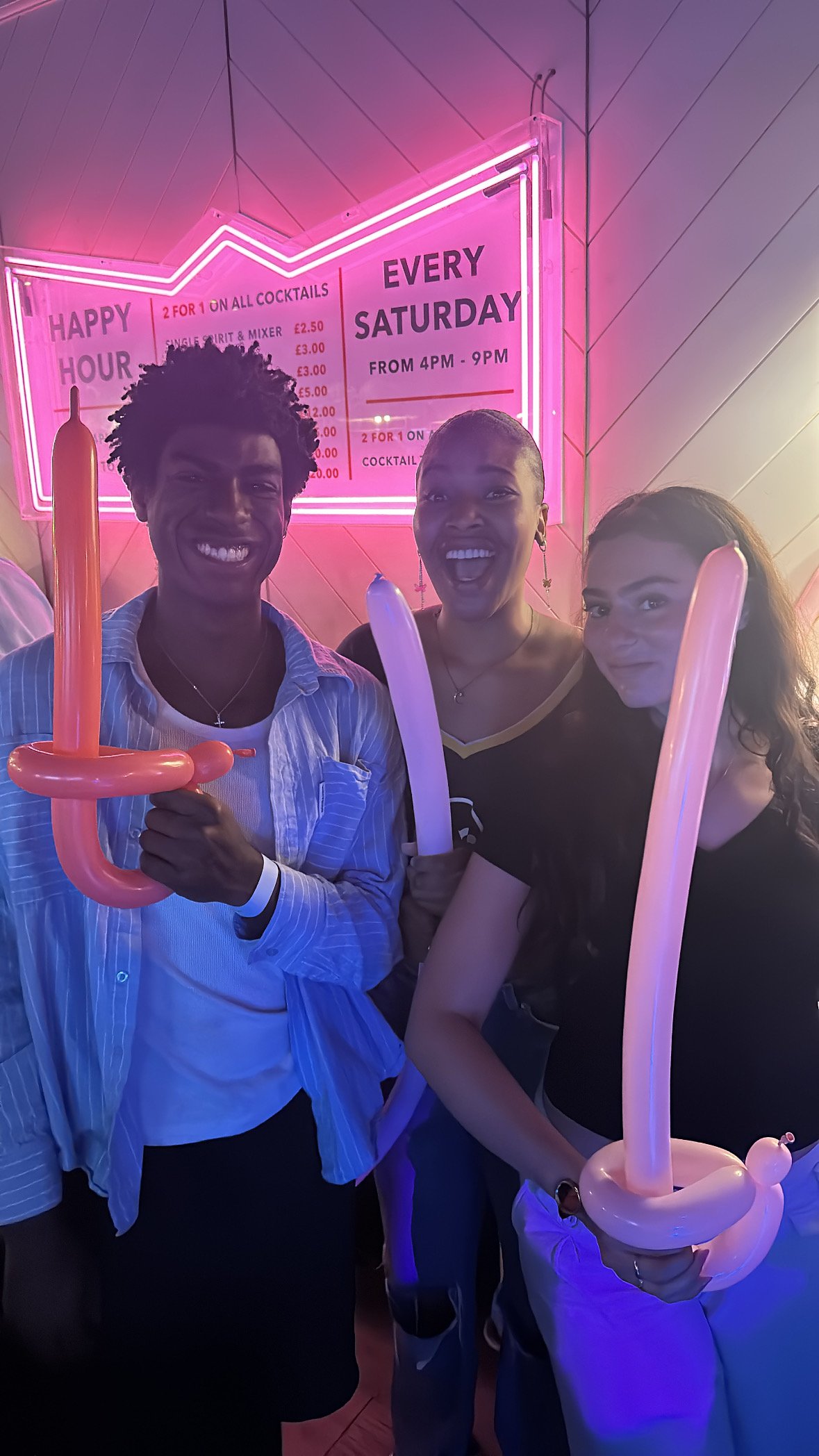 Three people smiling and holding balloon swords in front of a neon sign advertising happy hour and Saturday specials.