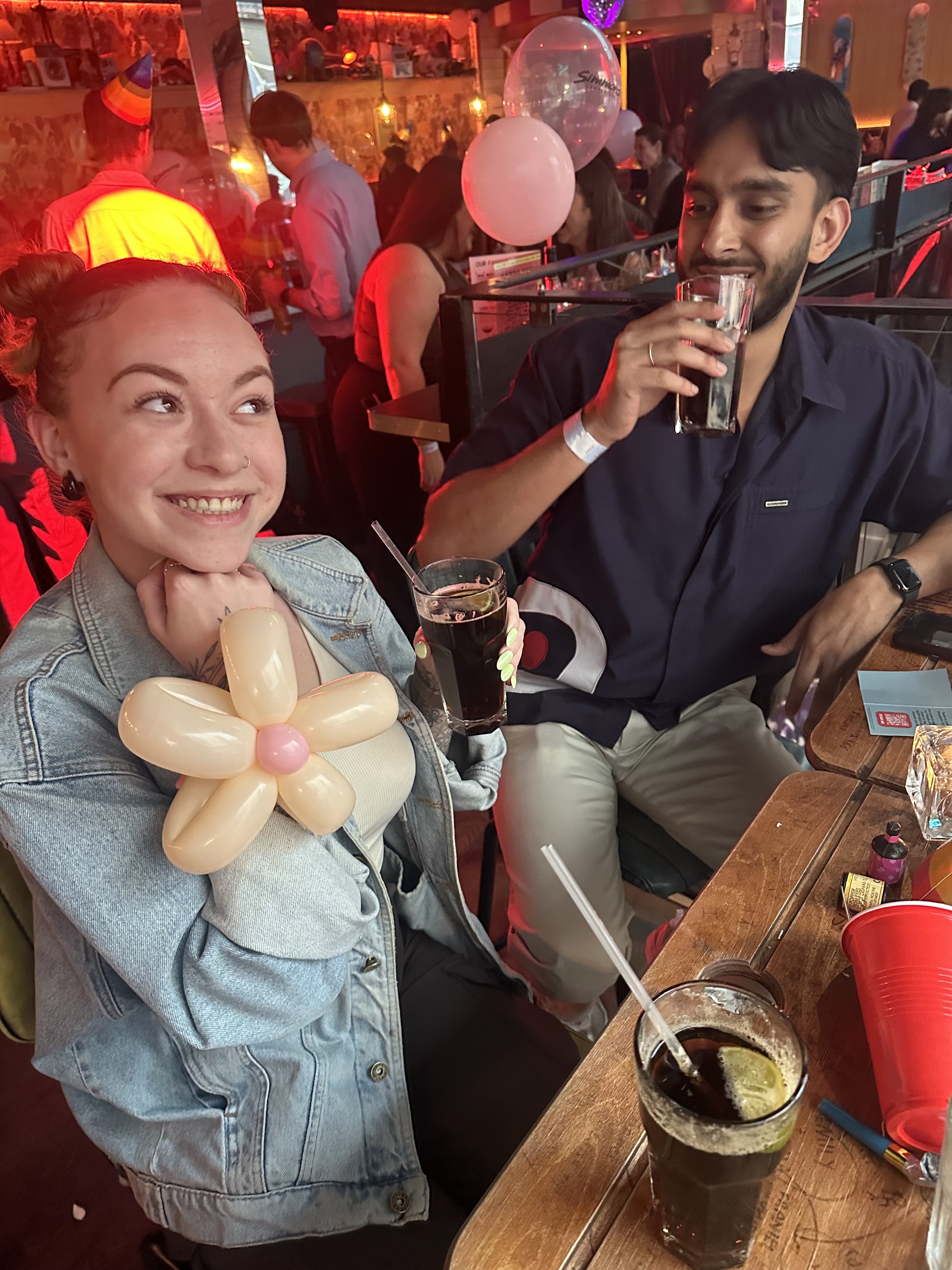 Two people sitting at a bar, smiling and holding drinks, with a balloon flower and colorful party decorations in the background.
