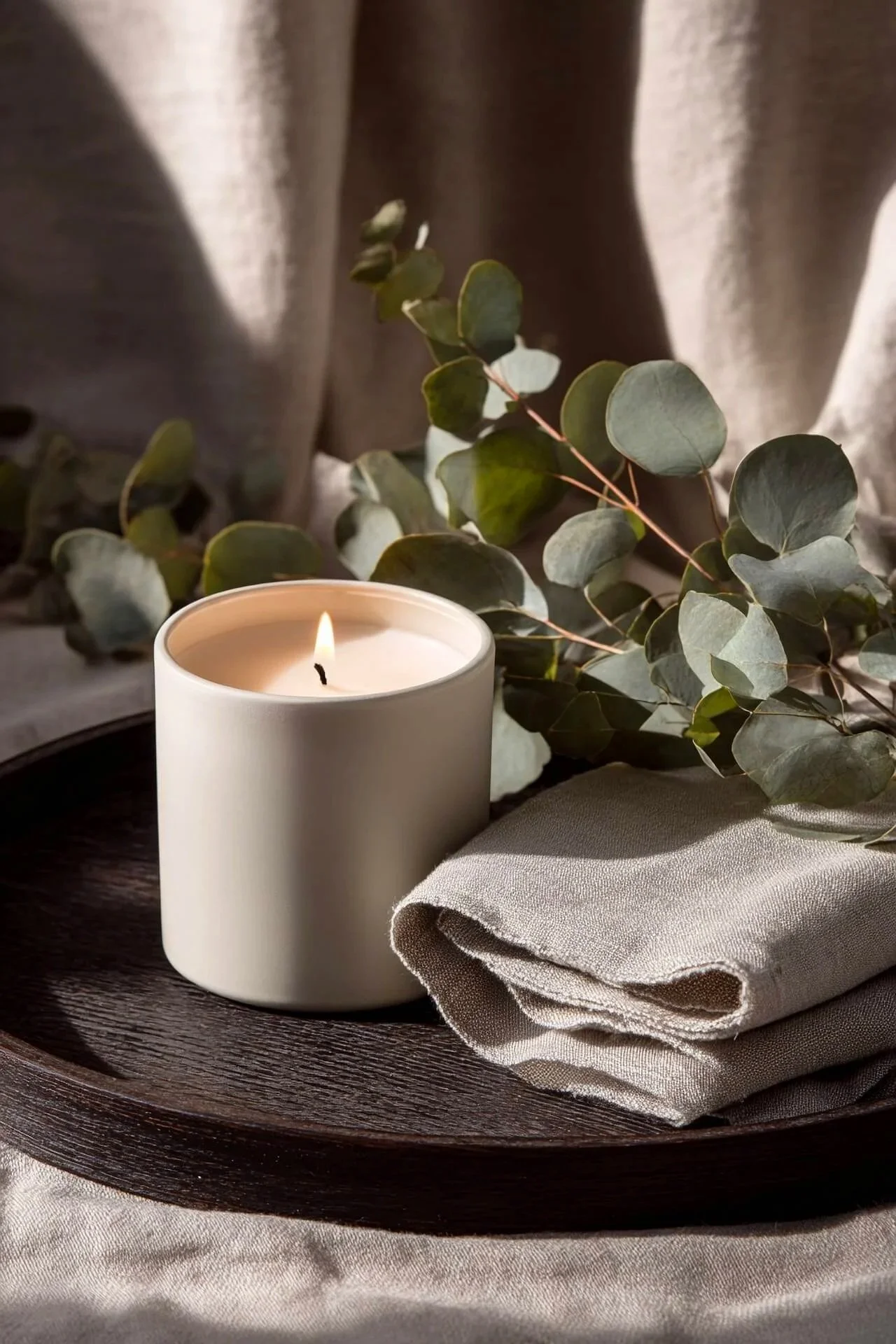 A lit candle on a brown tray with folded beige fabric to the right and  eucalyptus leaves resting on top
