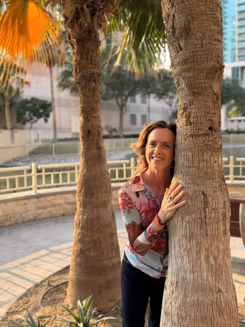 Michela Modolo standing hugging a tree trunk, smiling at the camera, with water and tree fronds in the background