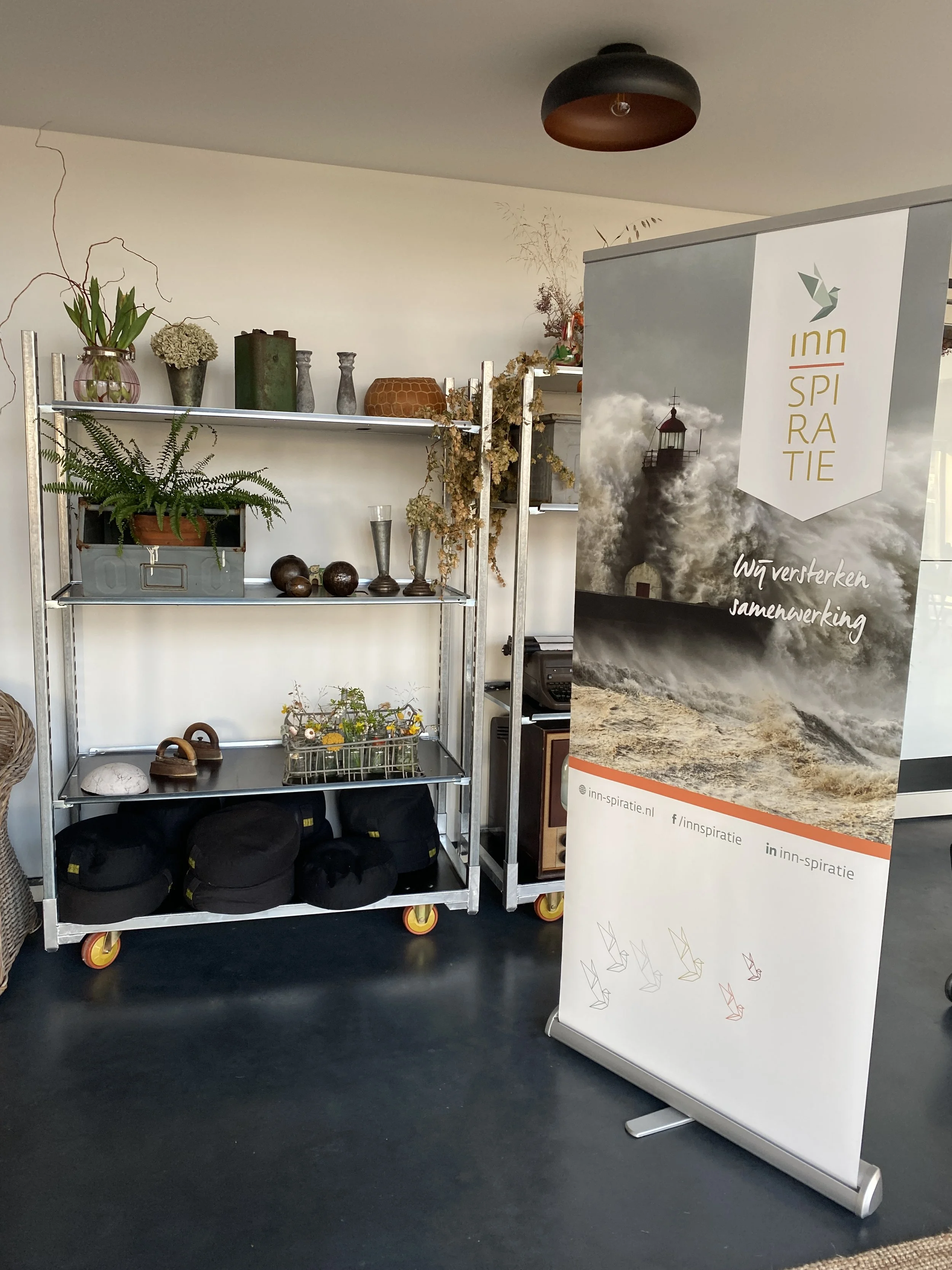 Interior of a room with a metal shelving unit holding decorative vases and plants. A large banner with an image of a lighthouse in stormy seas and the text 'Inn Spira Tie' and 'Wij versterken samenwerking' stands beside the shelves.