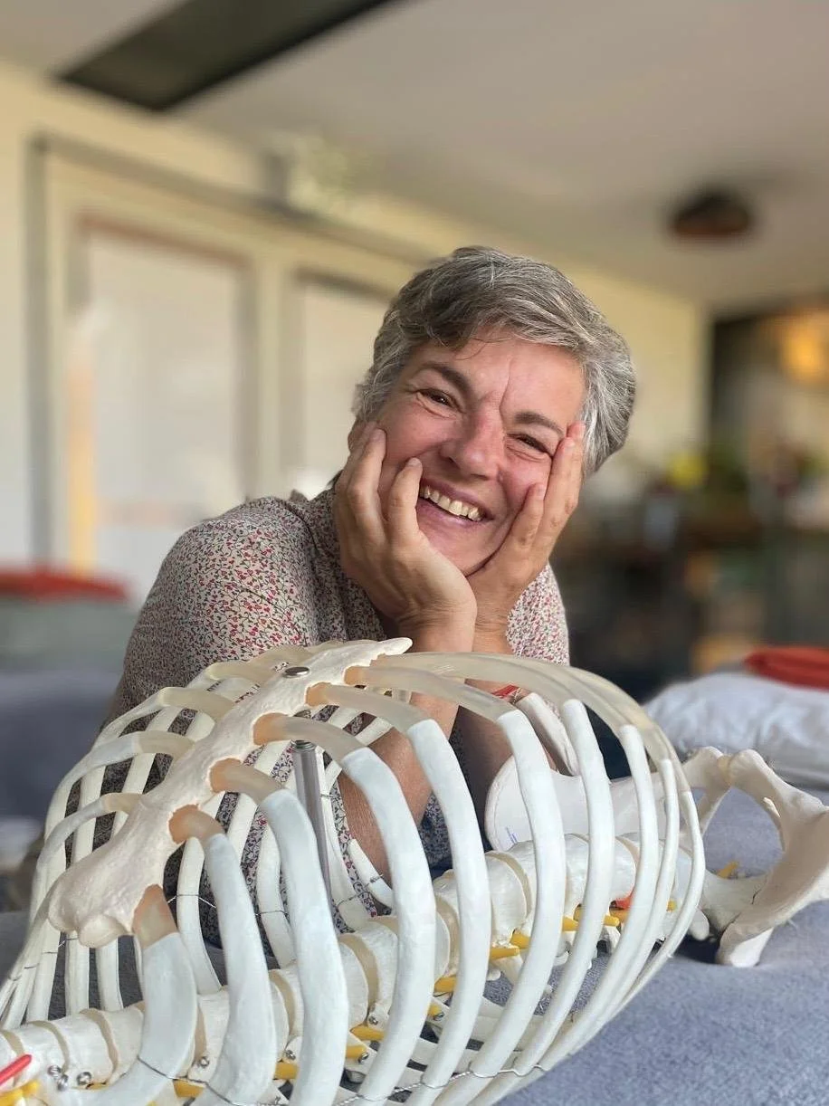 A smiling woman with short gray hair holding her face with both hands, sitting behind a plastic model of a human rib cage.