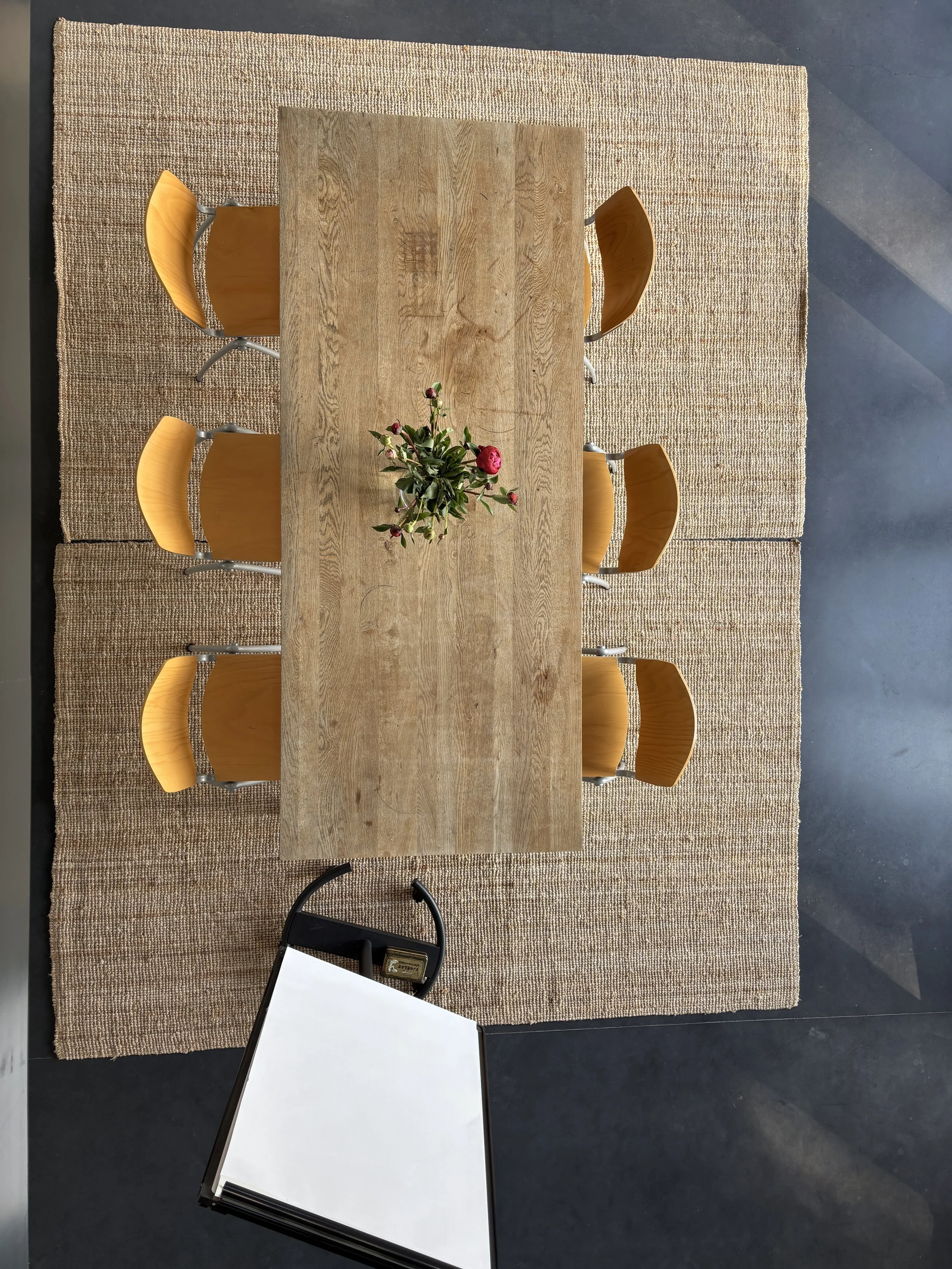 Top-down view of a wooden conference table with six yellow chairs, a flower arrangement in the center, on a woven rug with a whiteboard or flip chart at the bottom.