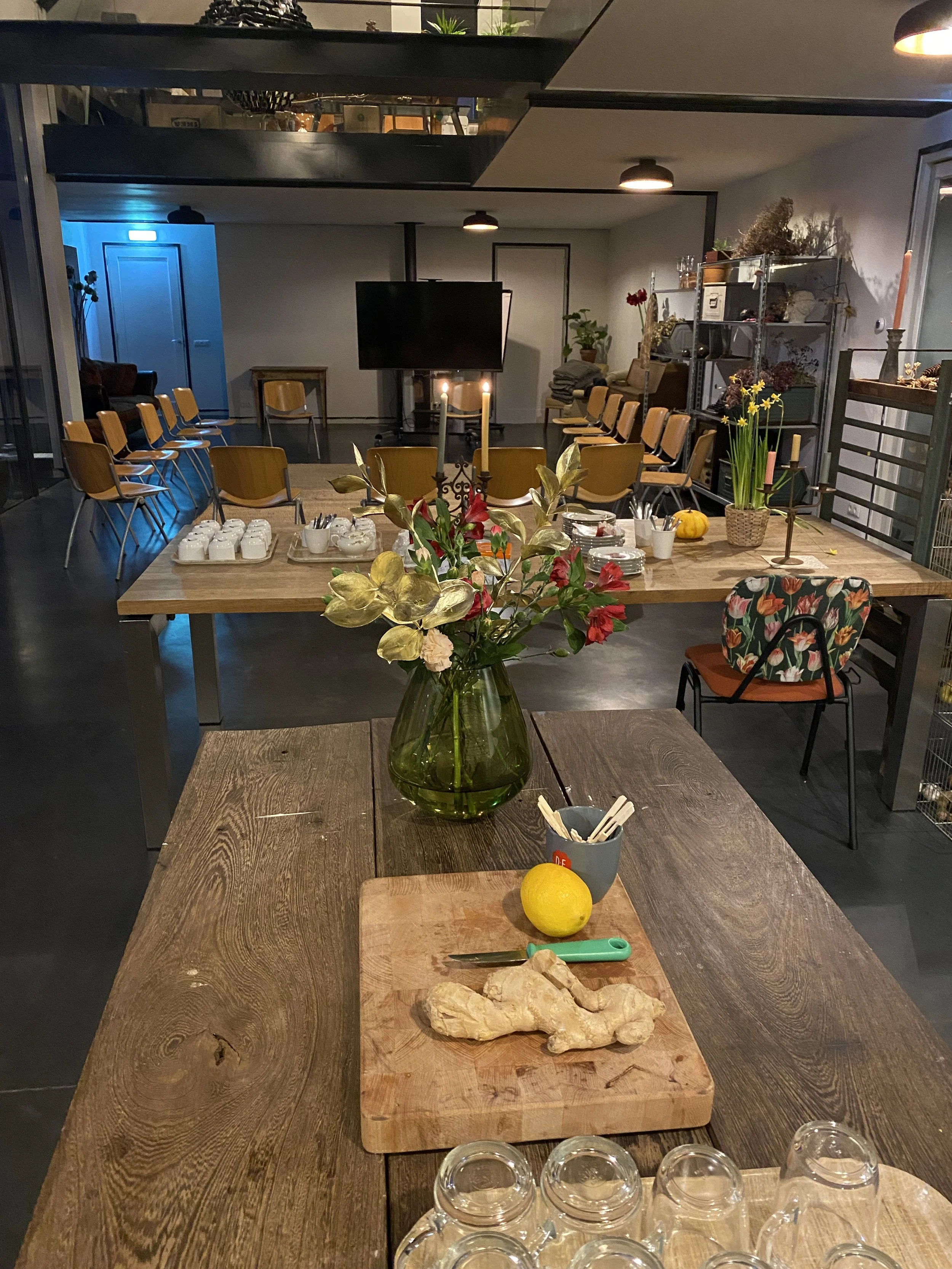 A dining area set up for a gathering in a modern, well-lit room. There is a large wooden table in the foreground with a vase of flowers, a lemon, a piece of ginger, and a cutting board. Behind it is another table with dishes, utensils, and decoration
