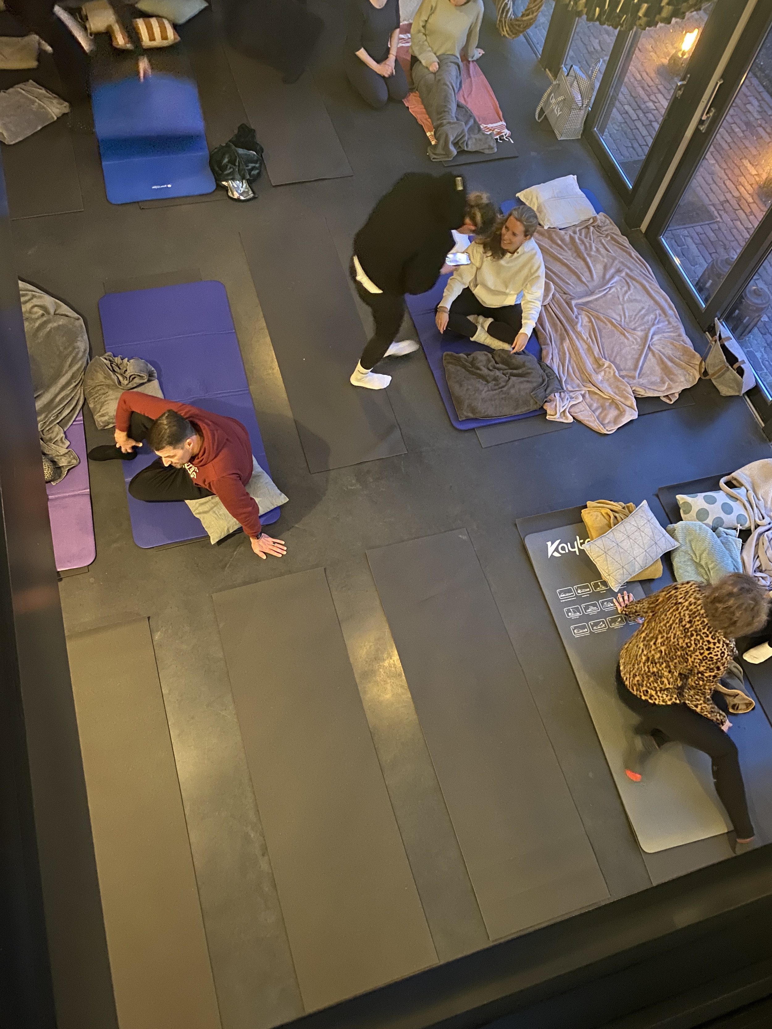 People participating in a yoga or meditation class in a studio with mats, blankets, and pillows. Some are seated or lying down, and there is a woman in a leopard print top and black pants at the front. An instructor is speaking to a seated woman near