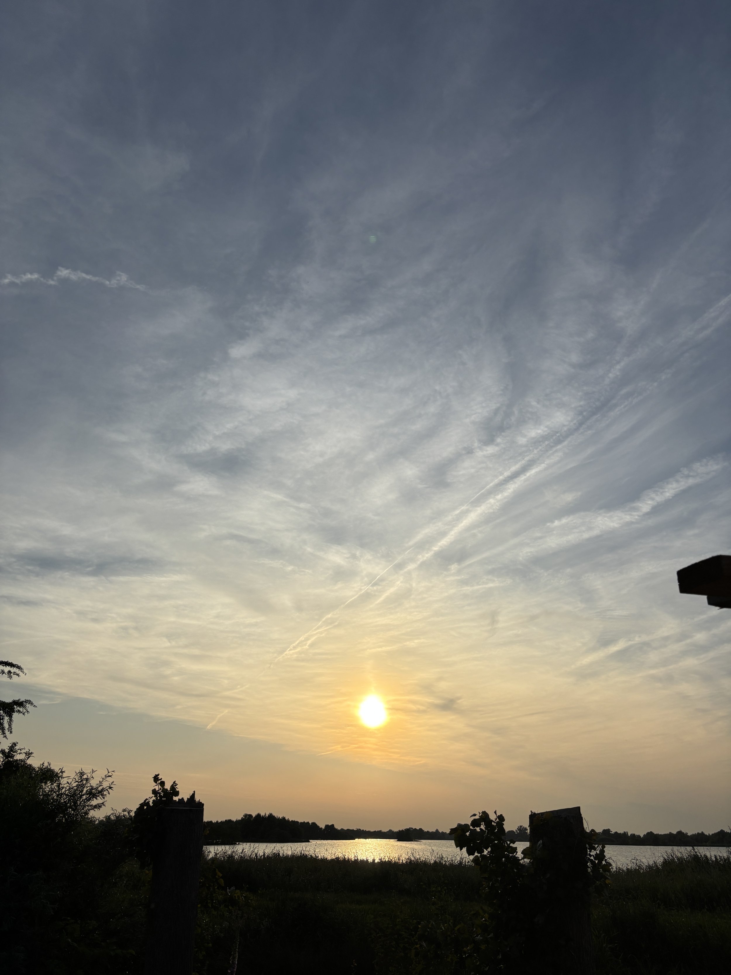 Sunset over a lake with silhouetted trees and bushes in the foreground, and a sky filled with wispy clouds.