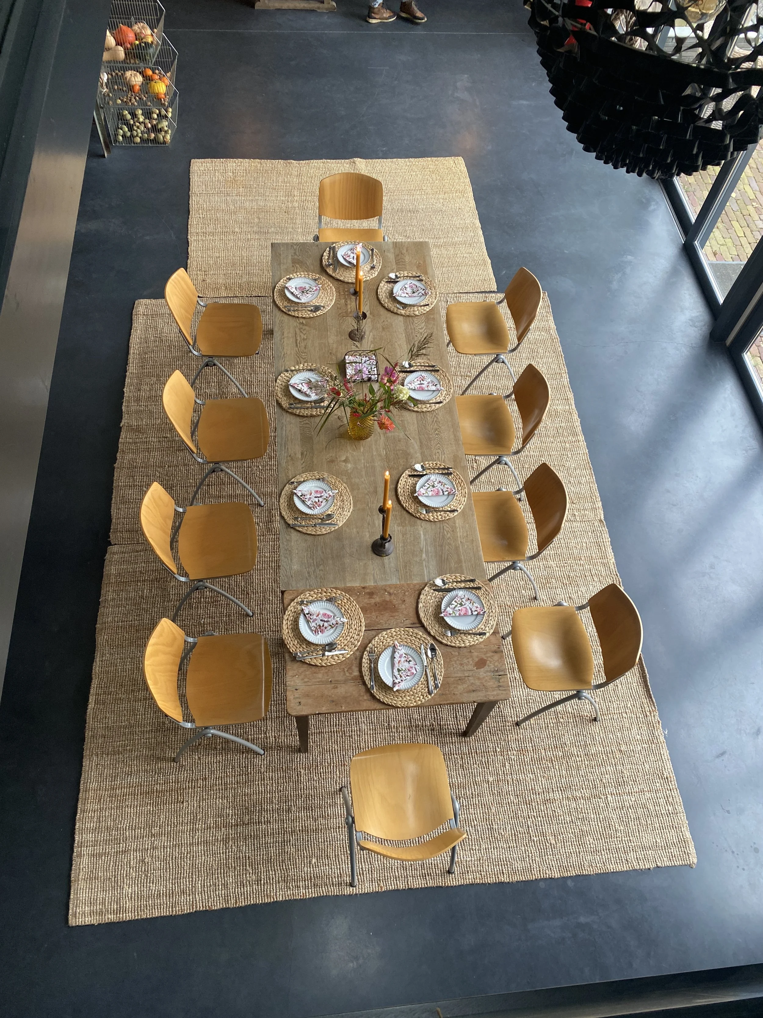 A dining table set for ten people with plates, napkins, and cutlery, decorated with a flower arrangement and two candles, on a woven rug, taken from an overhead perspective in a modern interior.