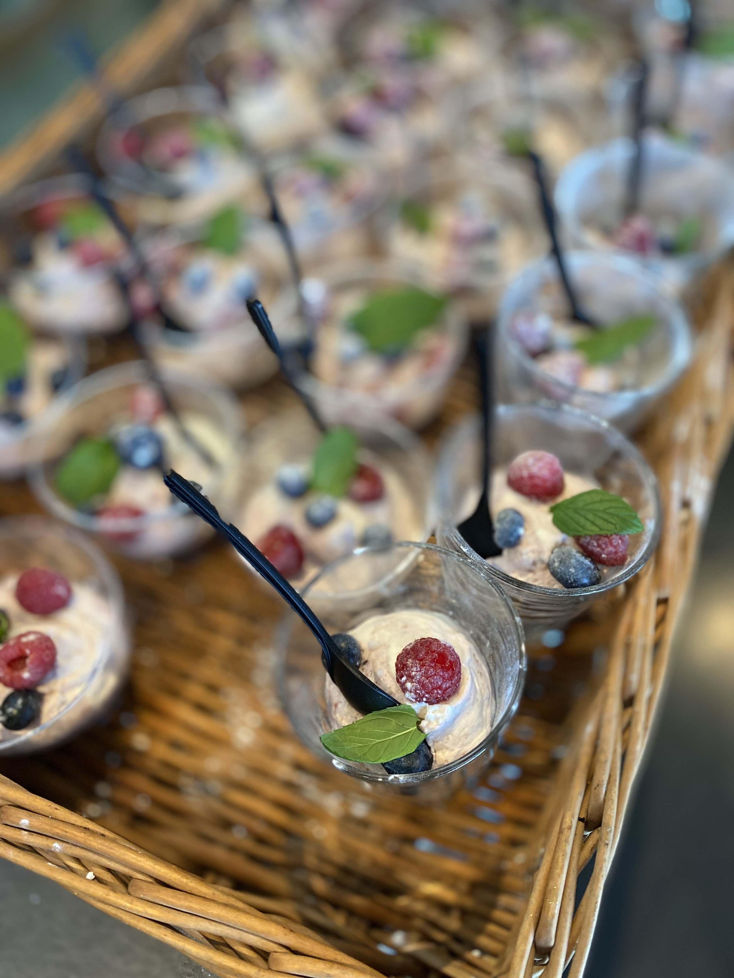 Several small glasses of creamy dessert topped with mixed berries and mint leaves on a woven tray.