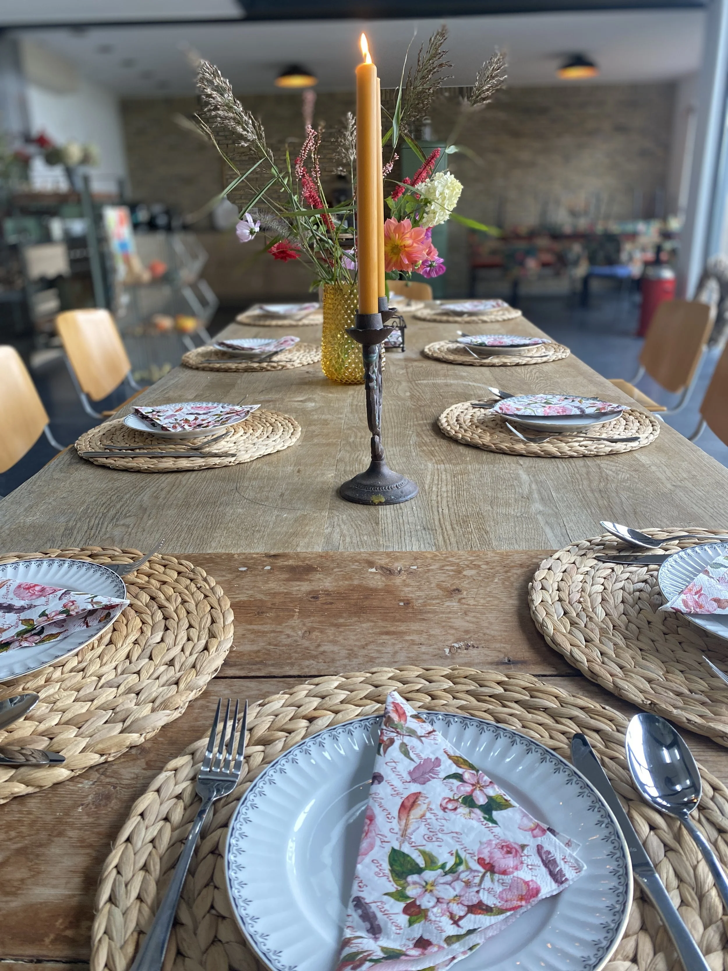 A long wooden dining table set for a meal with woven placemats, floral napkins, silverware, and a centerpiece with a candle and flowers, in a brightly lit room.