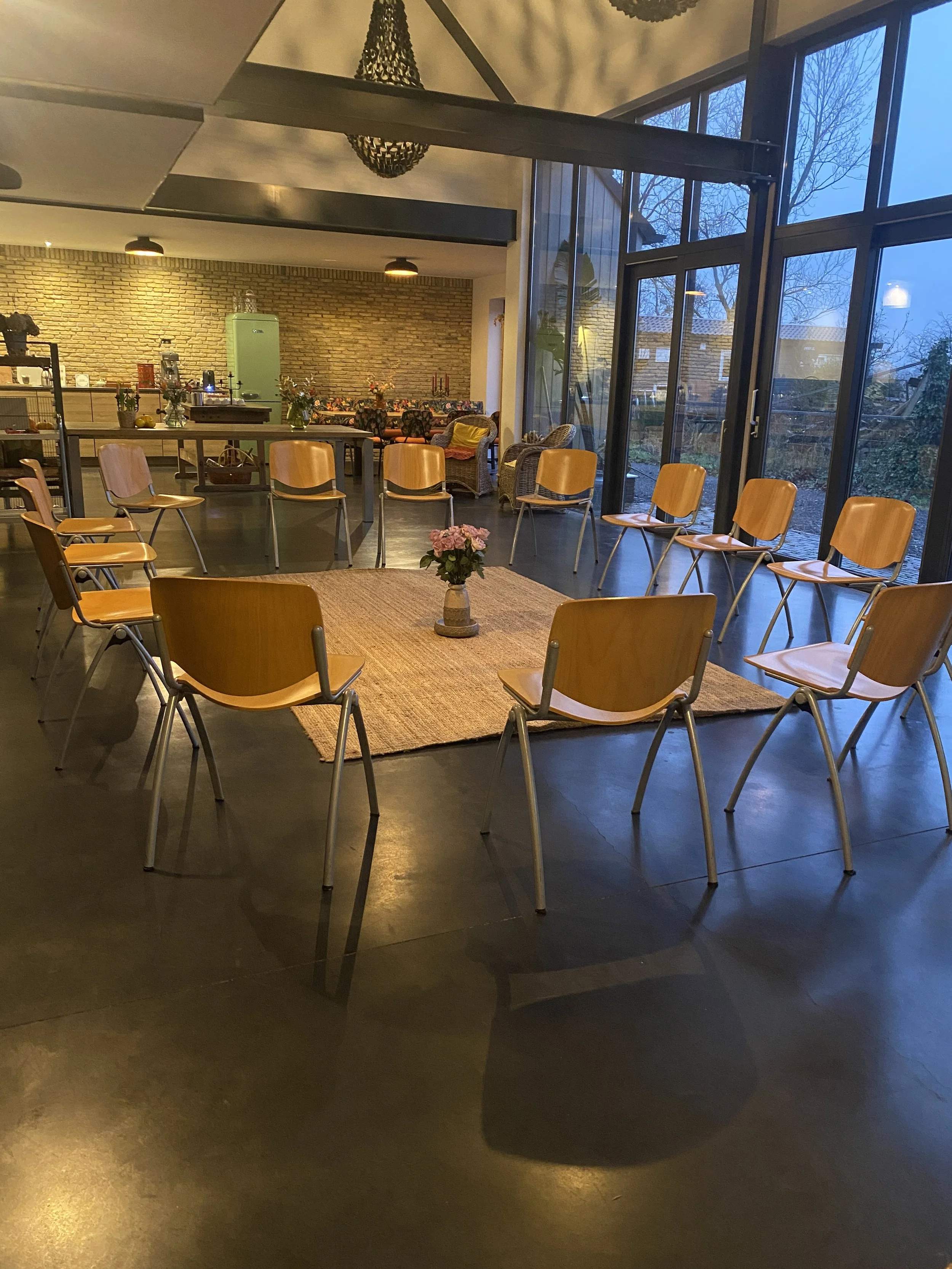 A spacious cafe or community space with a circle of wooden chairs around a small table with a pink flower arrangement. Large windows on the right let in natural light and show trees outside, and a brick wall is at the back with a buffet or serving ar