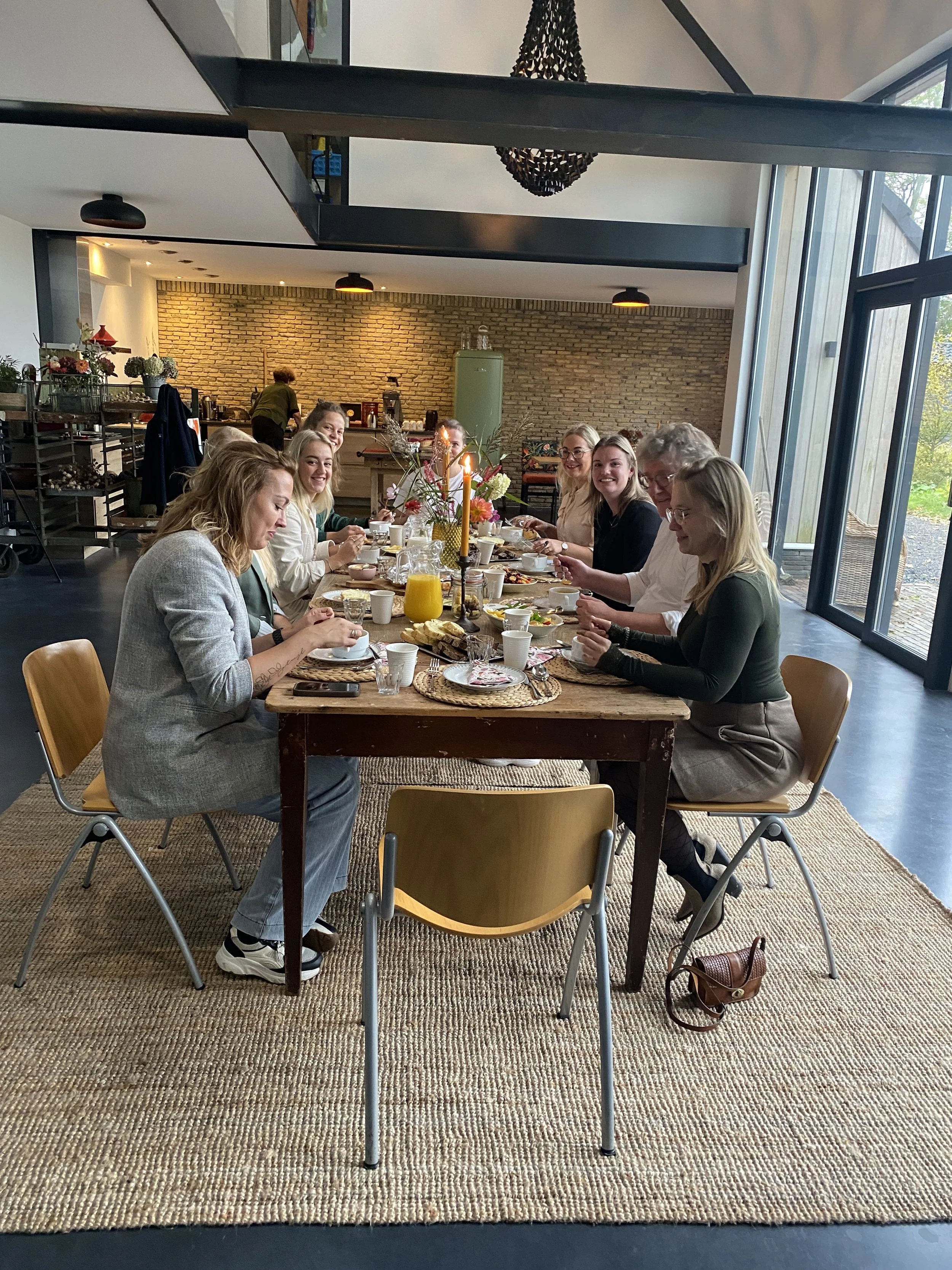 A group of nine people sitting at a long wooden table having a meal in a modern dining area with large window and brick wall in the background.