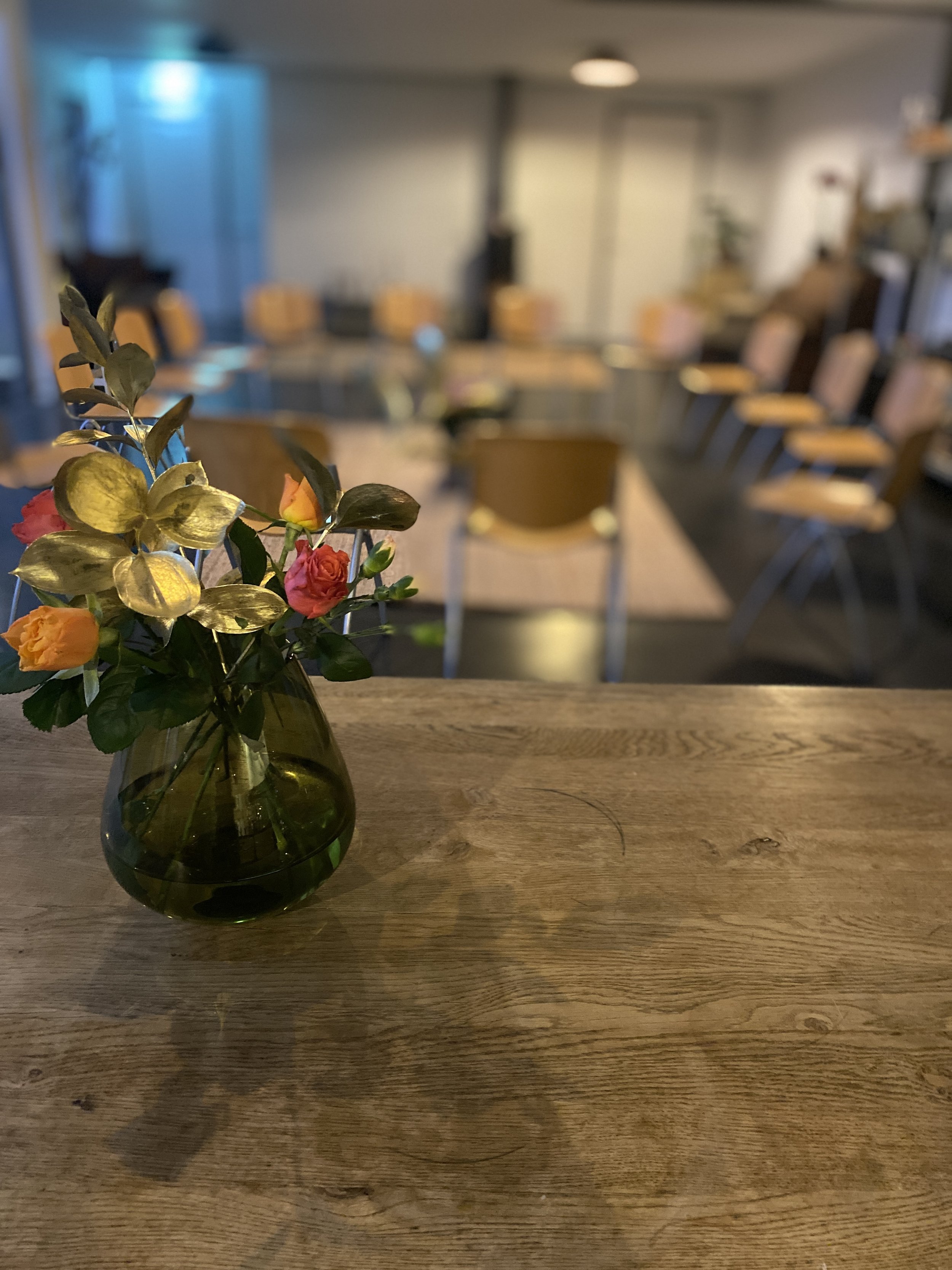 A small bouquet of colorful flowers in a green glass vase on a wooden table, with a blurred conference room in the background.