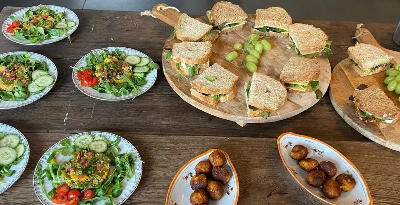 Assorted salads, sandwiches, and fried snacks on a wooden table. The salads include cucumbers, cherry tomatoes, and greens; the sandwiches contain lettuce and full grape bunches are on serving boards; the fried snacks are in small white dishes.