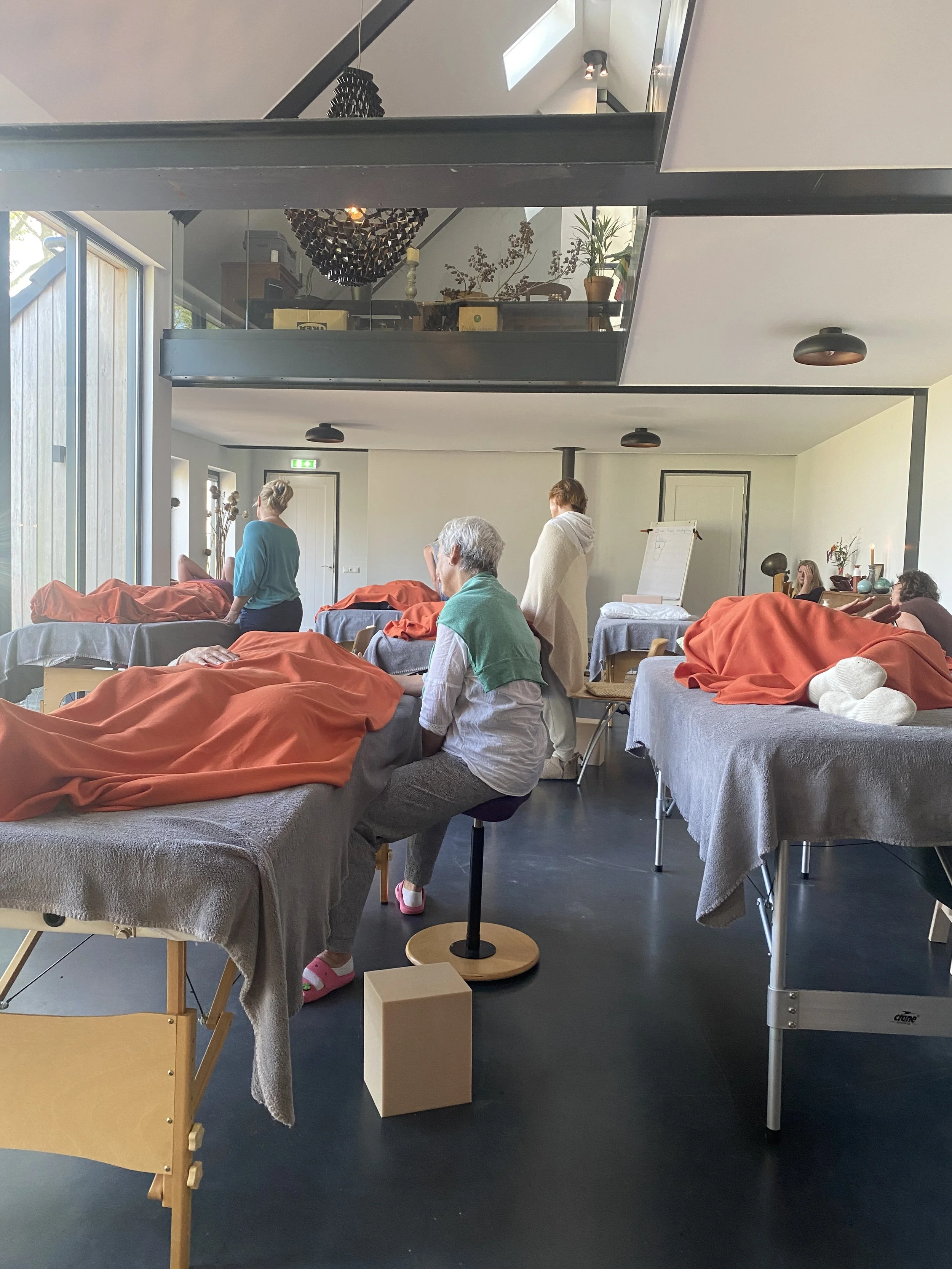 A room with several massage tables covered with orange and gray blankets, where people are receiving or waiting for massages. There are a few people sitting and standing, some with towels wrapped around them. The room has large glass windows, potted 