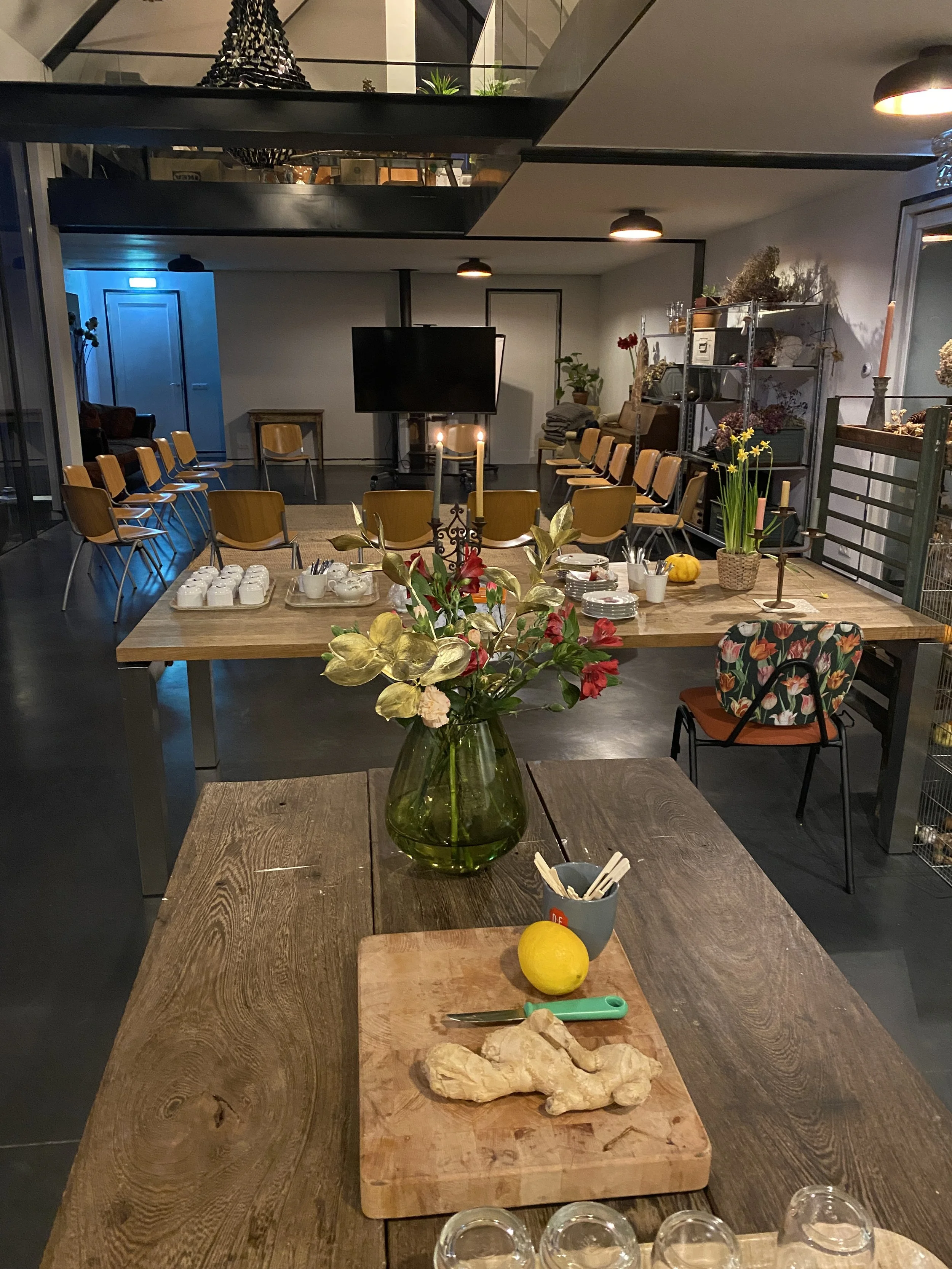 Interior of a room set up for a gathering with a long wooden table, a vase of flowers, and a cutting board with ginger, a lemon, and utensils, behind which are chairs arranged in a semi-circle with more chairs and decor on the walls.