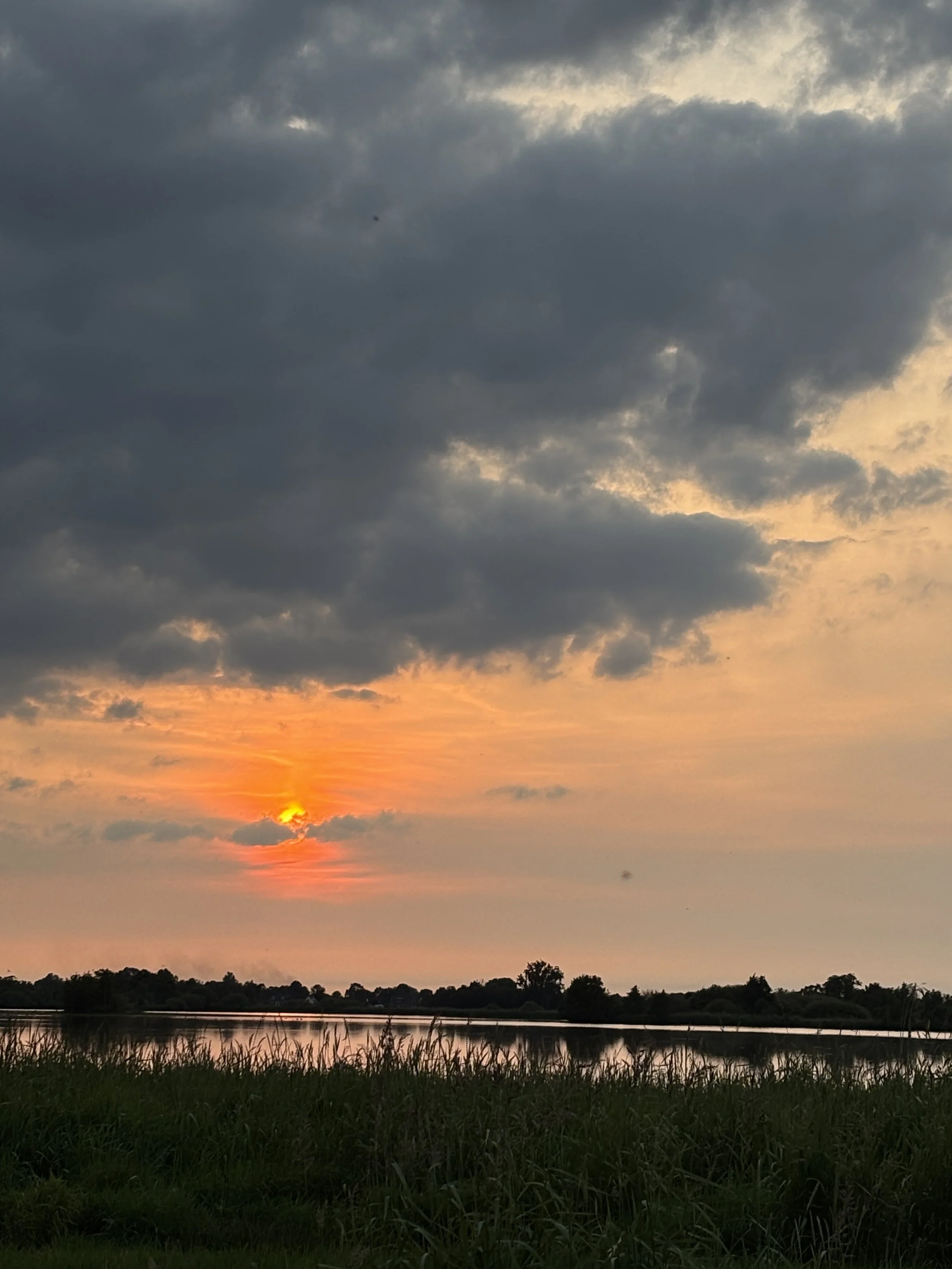 Sun setting over a body of water with dark clouds overhead and green grass in the foreground.