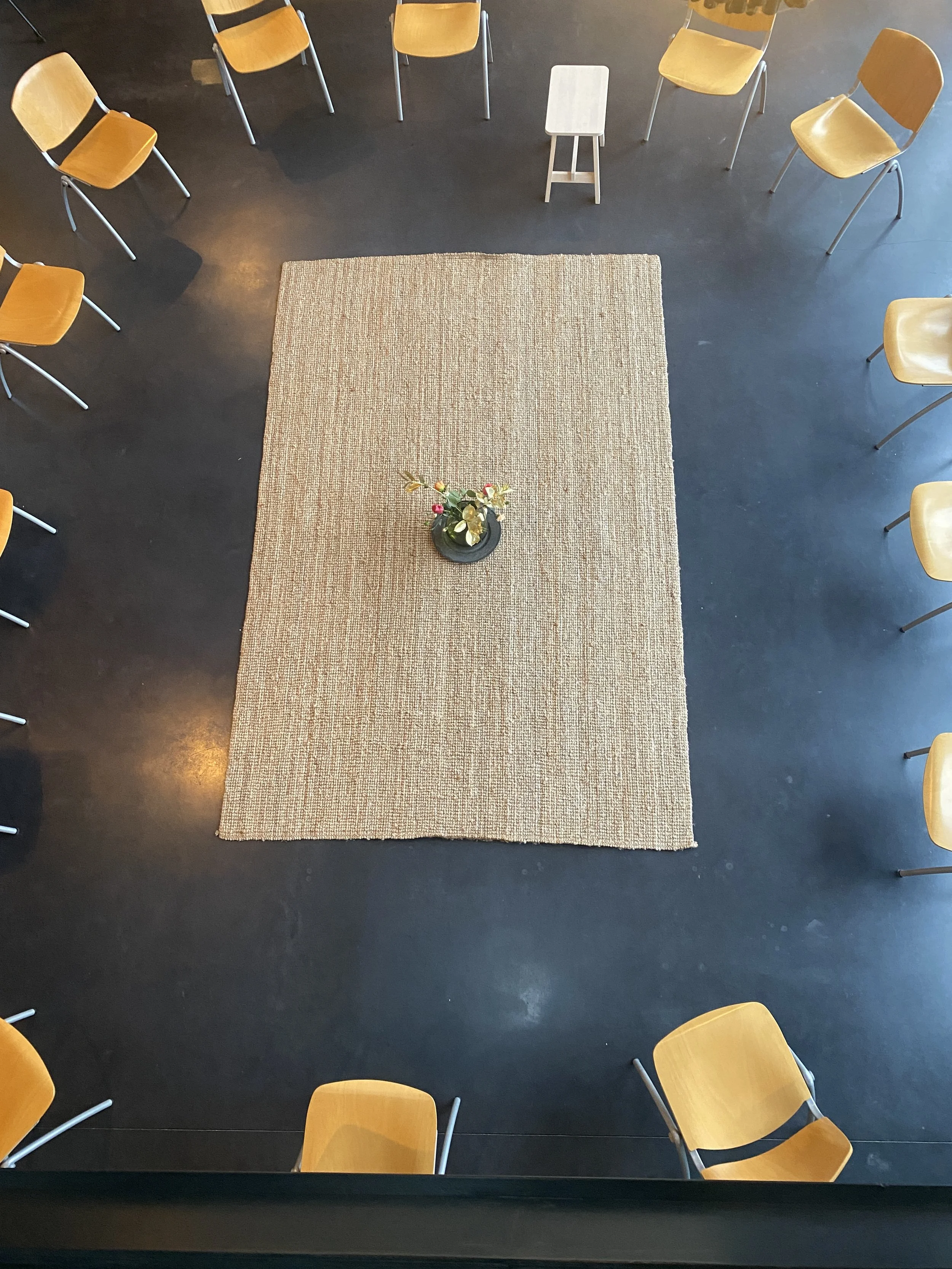 An overhead view of a beige rug with a flower arrangement in a black vase in the center surrounded by wooden chairs arranged in a semi-circle on a dark floor in a room.
