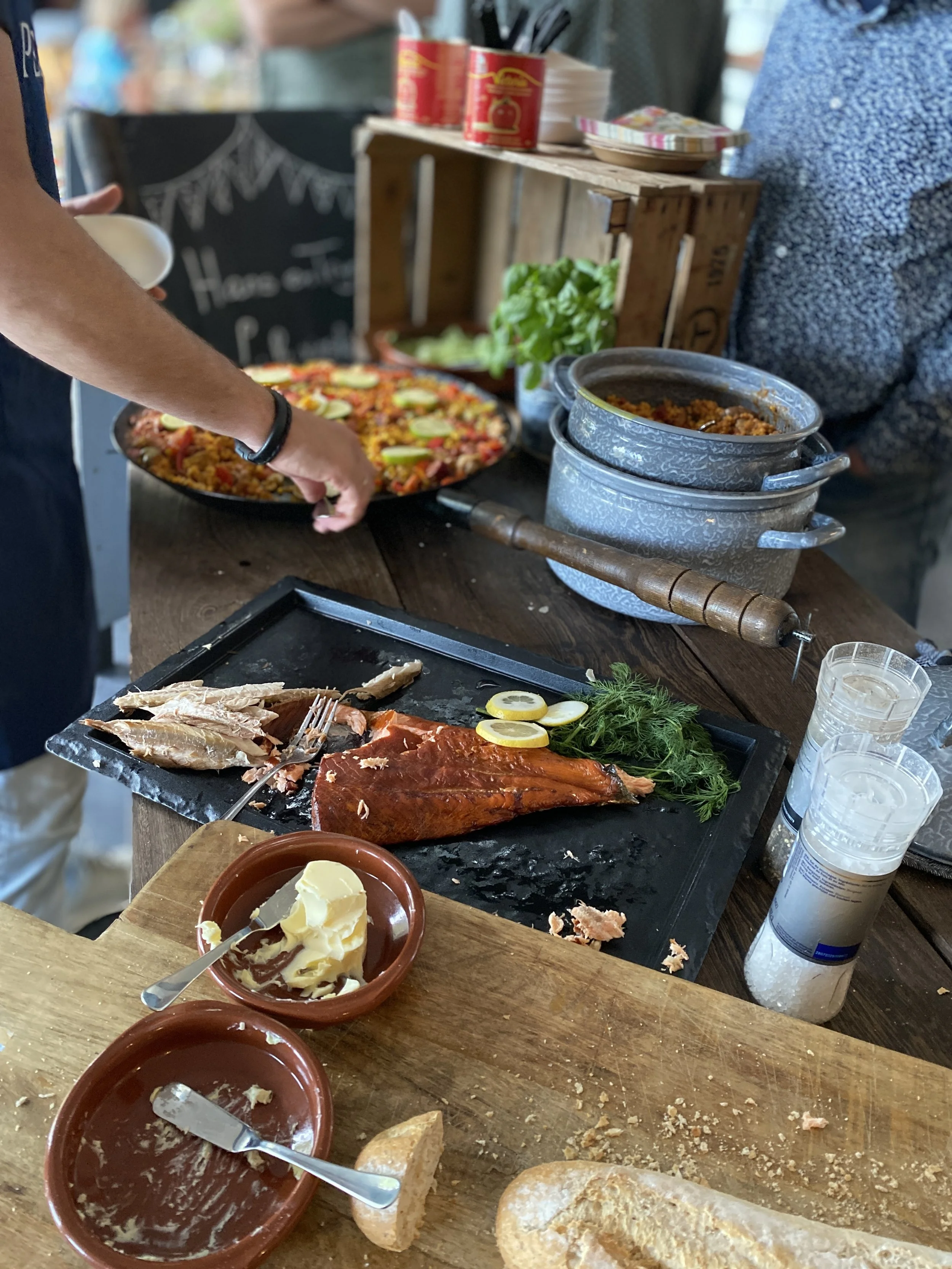 A table of food at a buffet with roasted meat, butter, bread, a black cast iron skillet with a rice dish topped with vegetables, and a pizza with slices of lemon and fresh herbs. There are containers of salt and pepper on the table.
