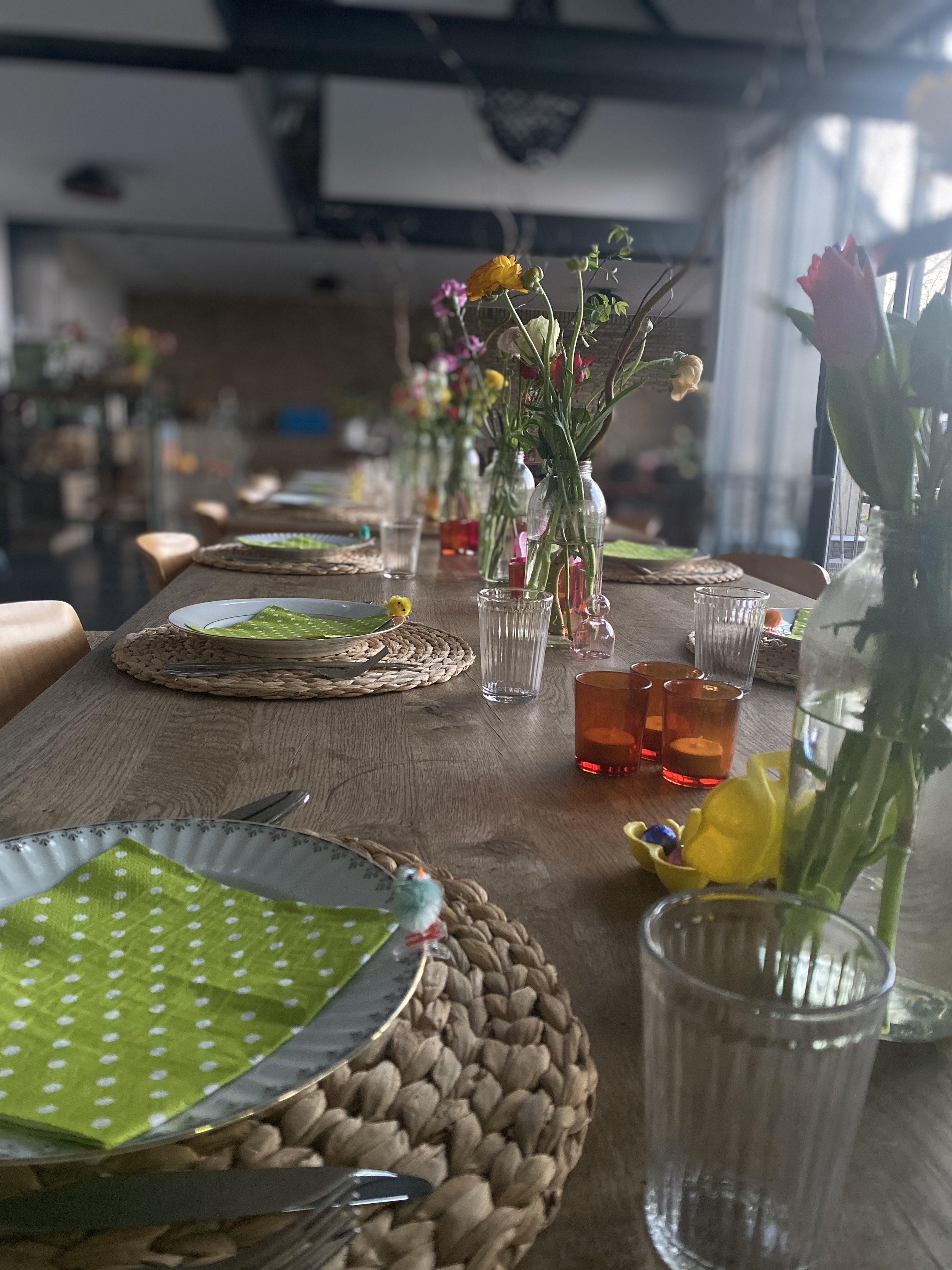 A long wooden dining table decorated with colorful flowers in glass jars, woven placemats, green polka dot napkins on white plates, and various glassware and candles, set in a room with large windows and natural light.