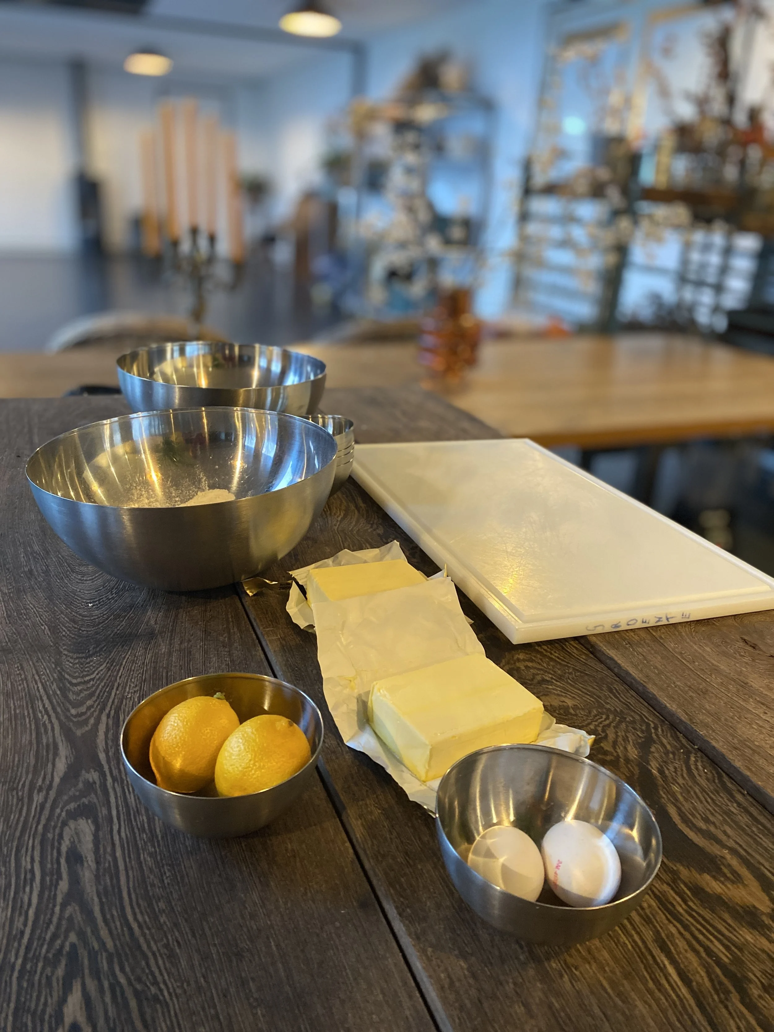 Kitchen counter with stainless steel mixing bowls, lemons, butter, eggs, a white cutting board, and parchment paper, with a blurred background of a dining or bakery space.