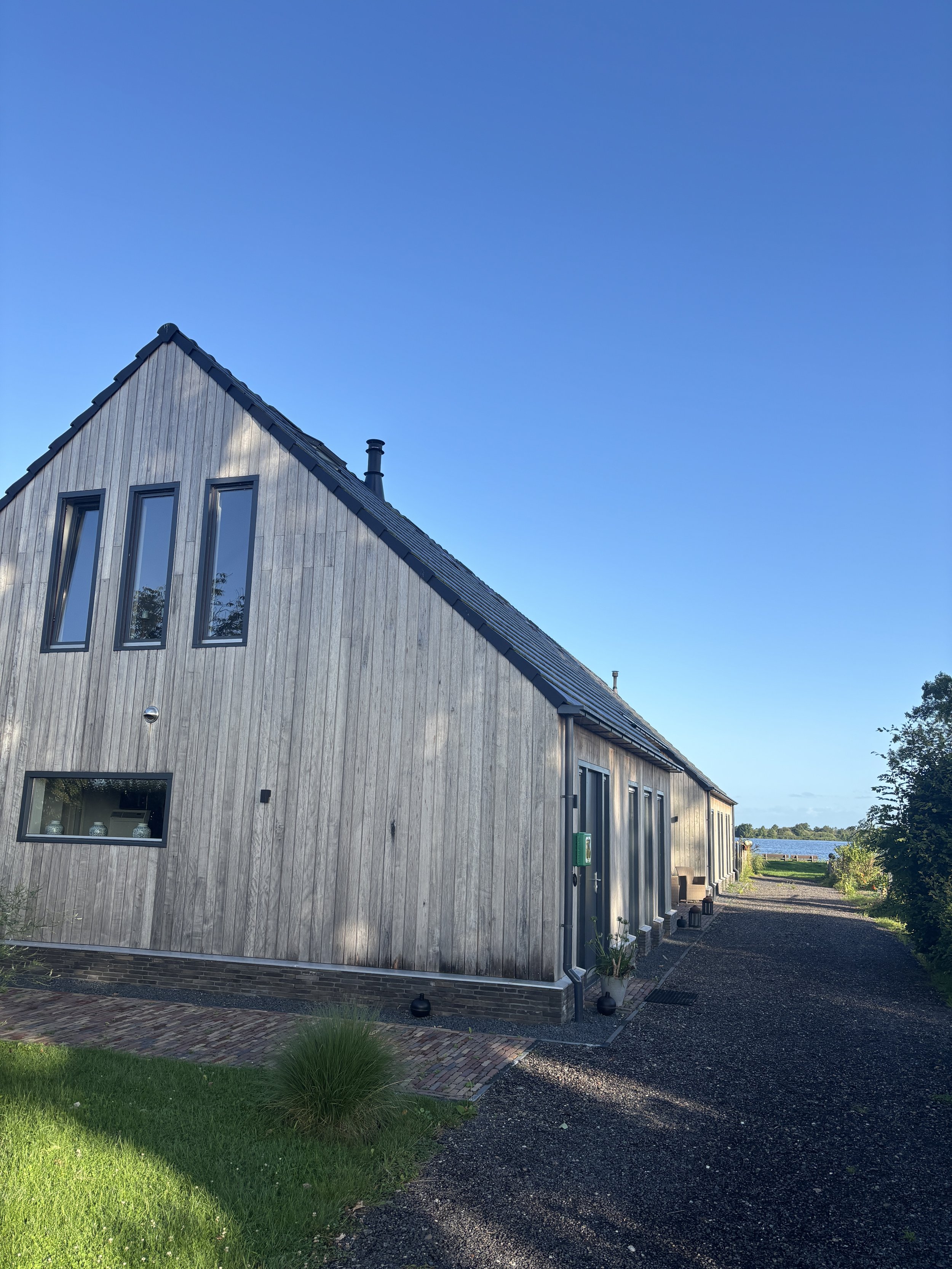 A modern, wooden house with a triangular roof, multiple windows, and a gravel pathway outside, near water and greenery under a clear blue sky.