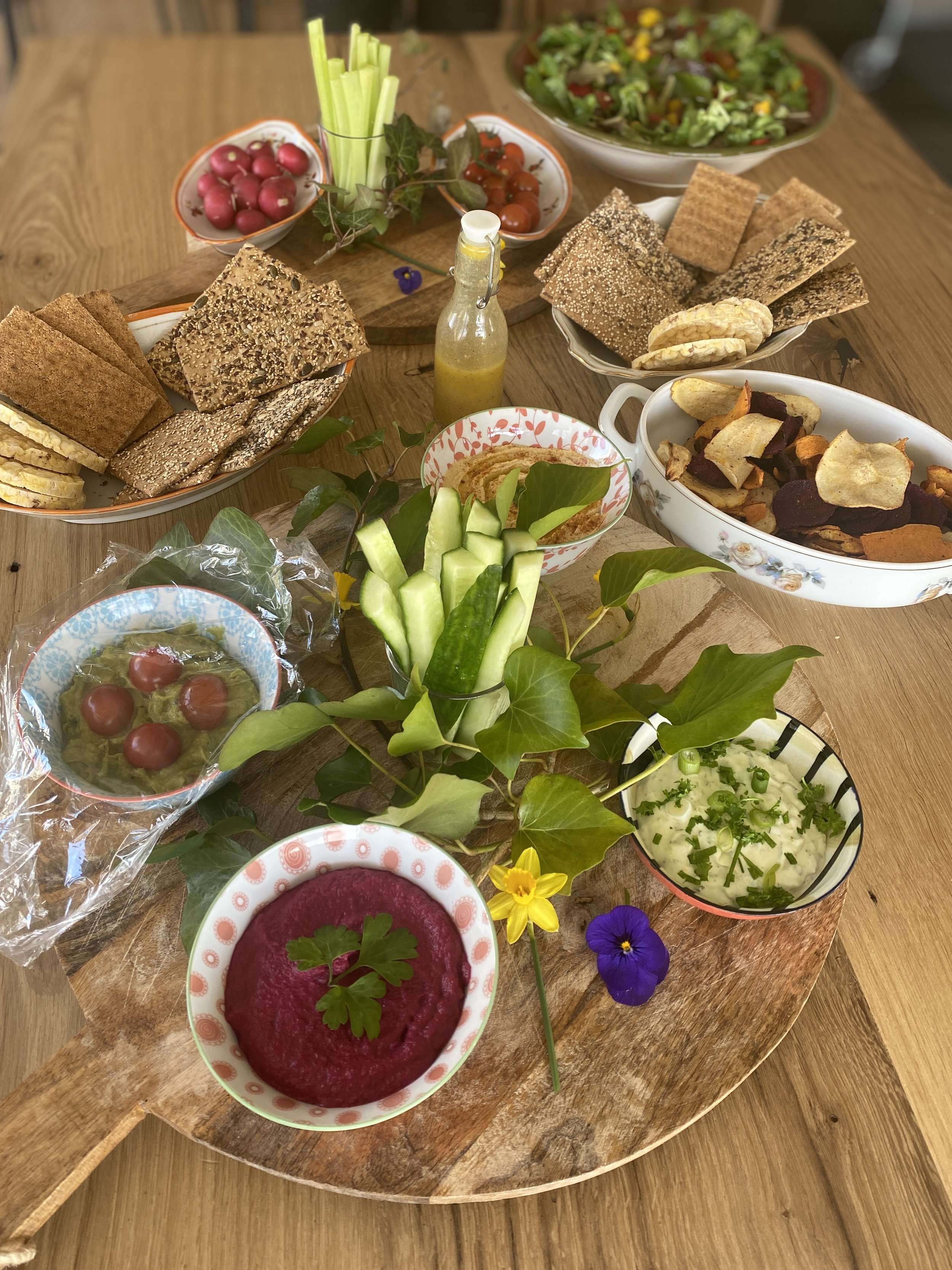 An assortment of appetizers and snacks on a wooden table, including sliced bread, veggie sticks, radishes, tomatoes, cucumber, hummus, beet dip, cherry tomatoes, and vegetable chips, decorated with edible flowers and green leaves.