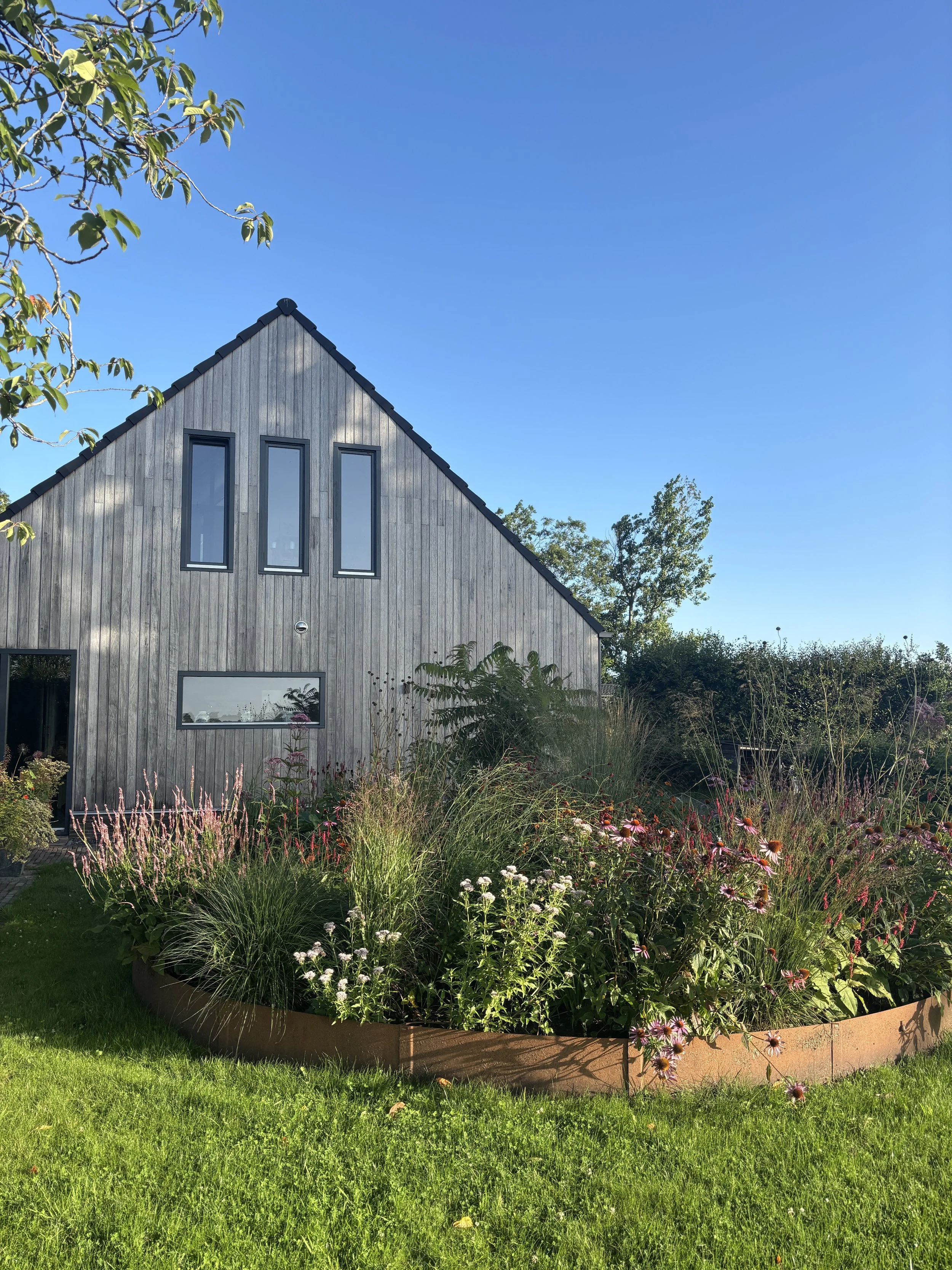 Modern house with wooden exterior and tall narrow windows, surrounded by a garden with various flowering plants and a green lawn under a clear blue sky.